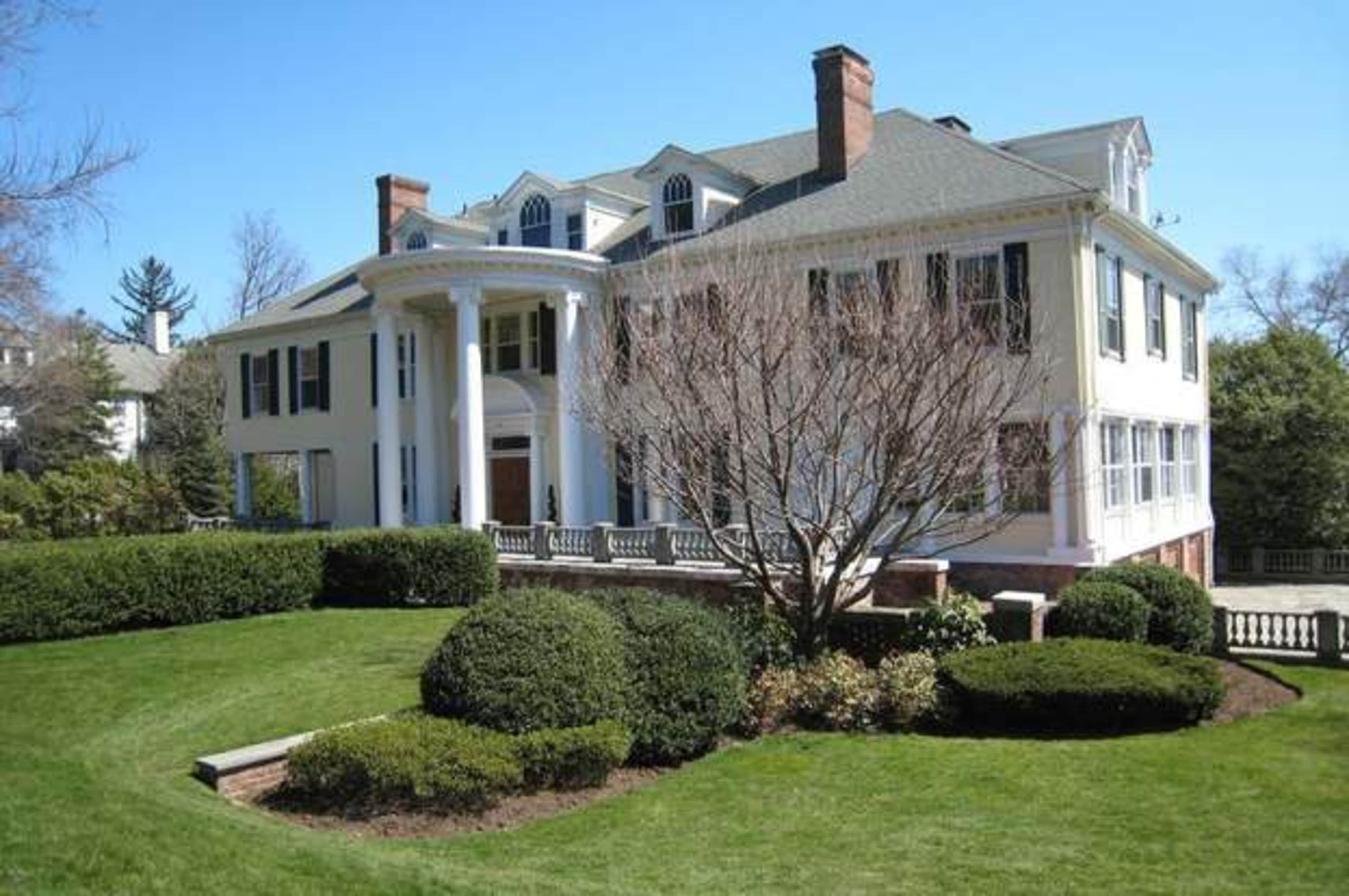 A large, elegant two-story house with white siding, columns at the entrance, and manicured lawns and shrubs surrounding the property.