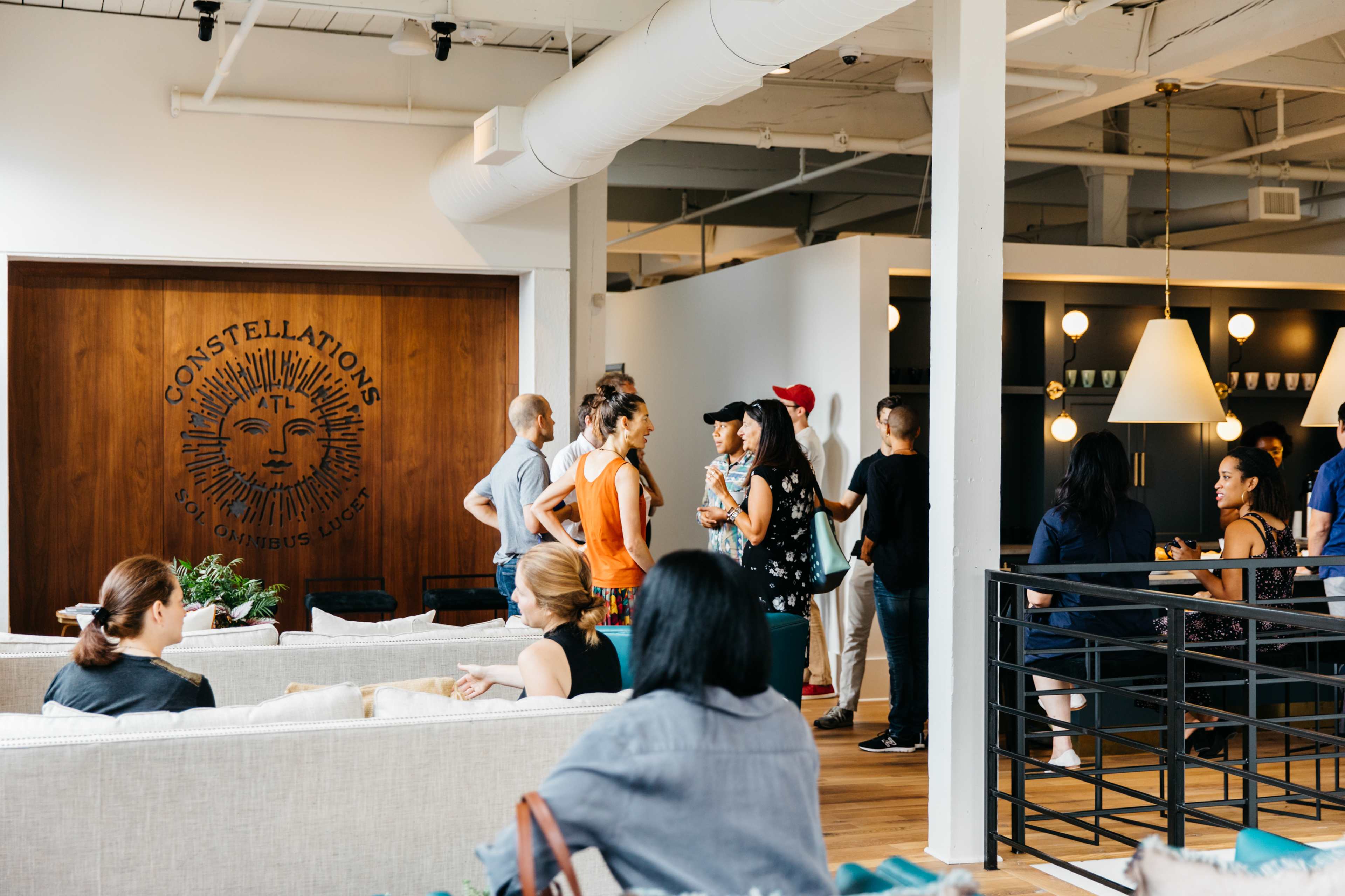 A group of people is gathered in a modern, open space featuring a decorative wall with the word "CONSTELLATIONS" and various seating arrangements.
