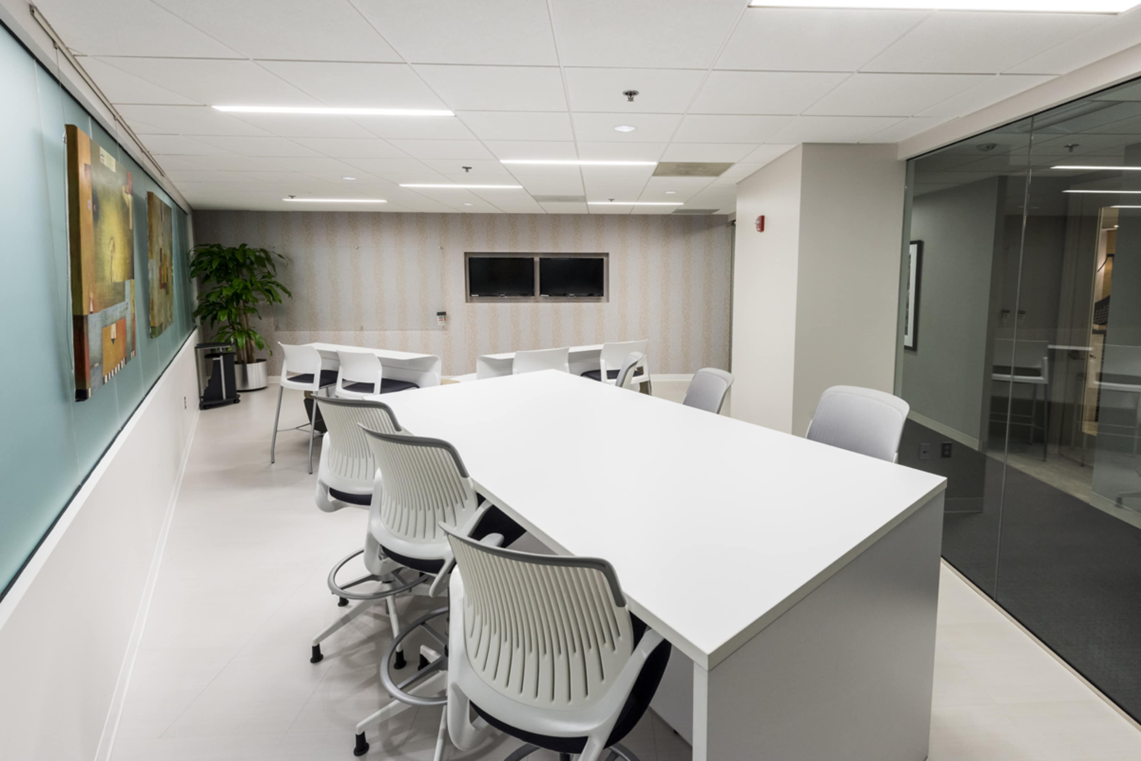 A modern conference room features a large white table surrounded by high-backed chairs, with a glass wall and a television display in the background.