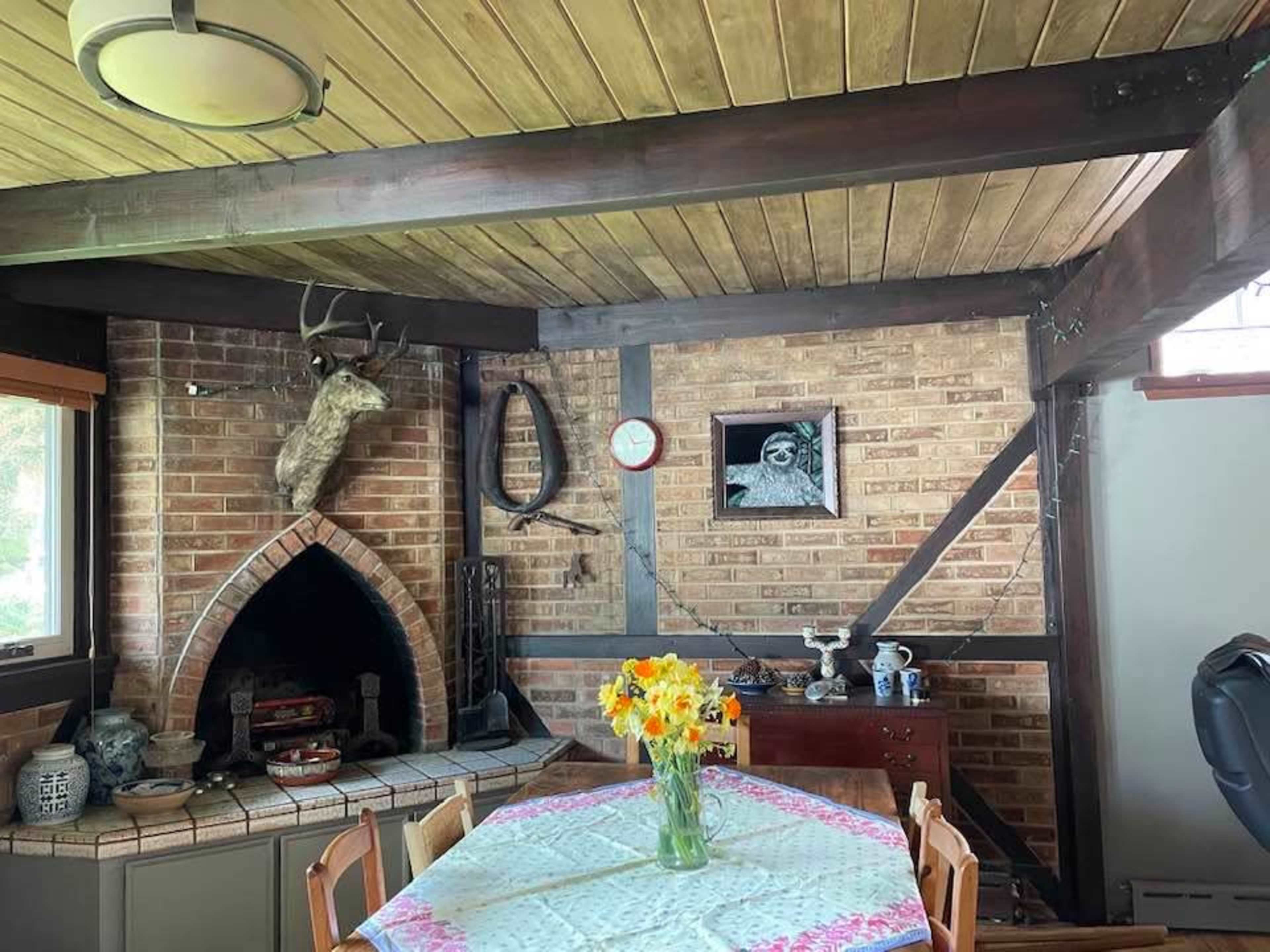 The interior features a wooden ceiling, a brick wall with a fireplace, a dining table adorned with flowers, and various wall decorations.