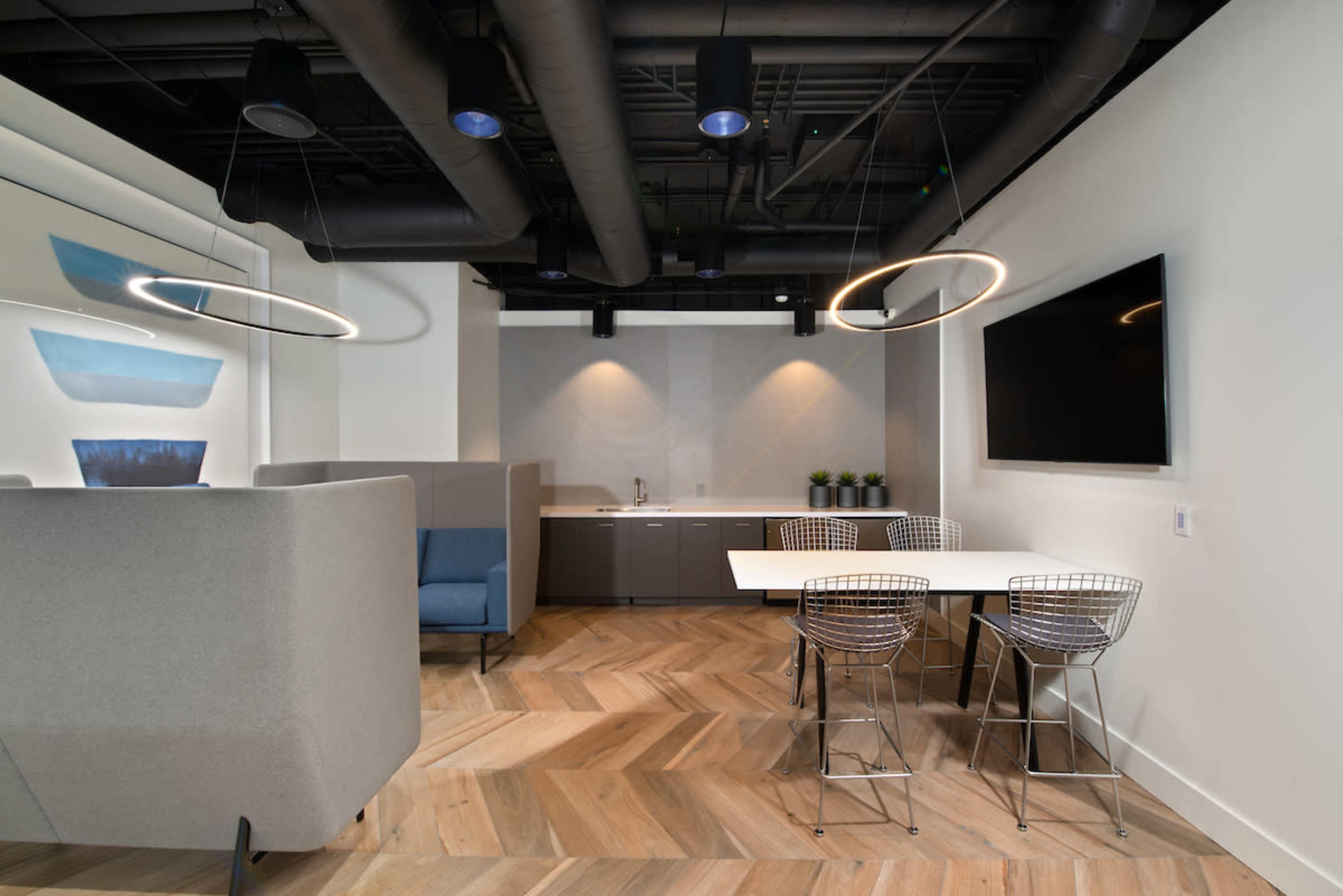 The image shows a modern office space featuring a kitchenette, a table with wire chairs, and soft seating areas under circular pendant lights.