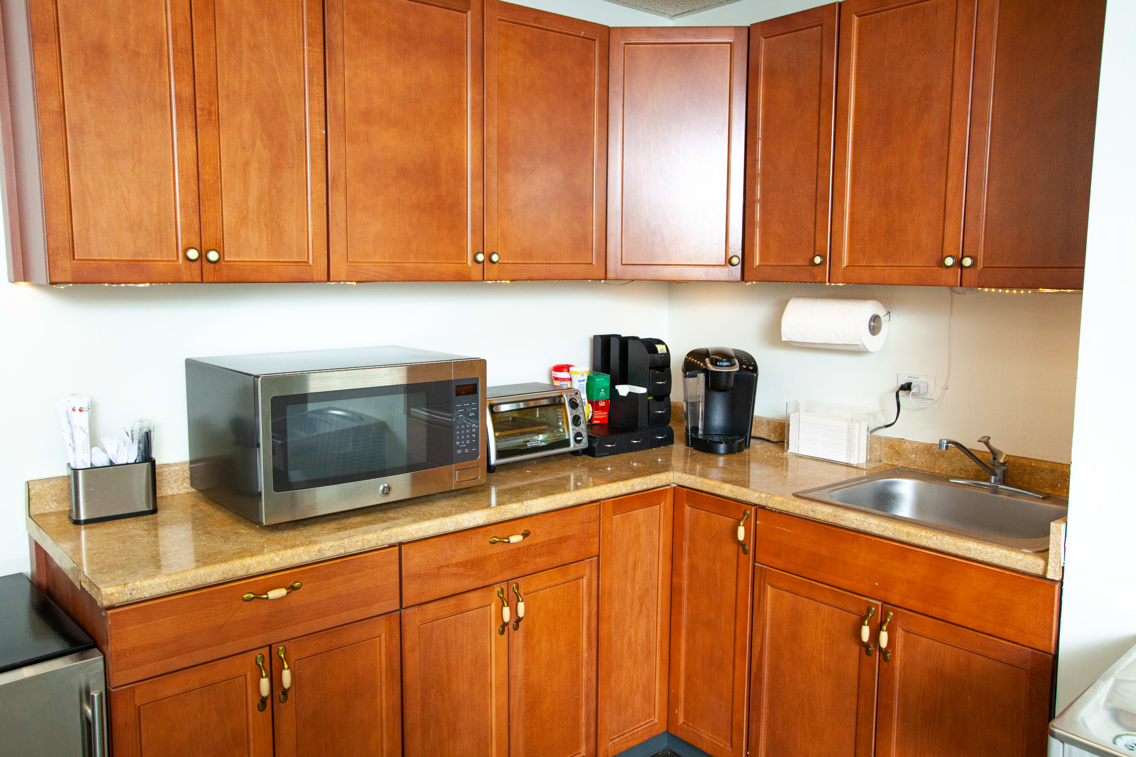 A kitchen corner featuring wooden cabinets, a microwave, a toaster, a coffee maker, and a small sink on a granite countertop.