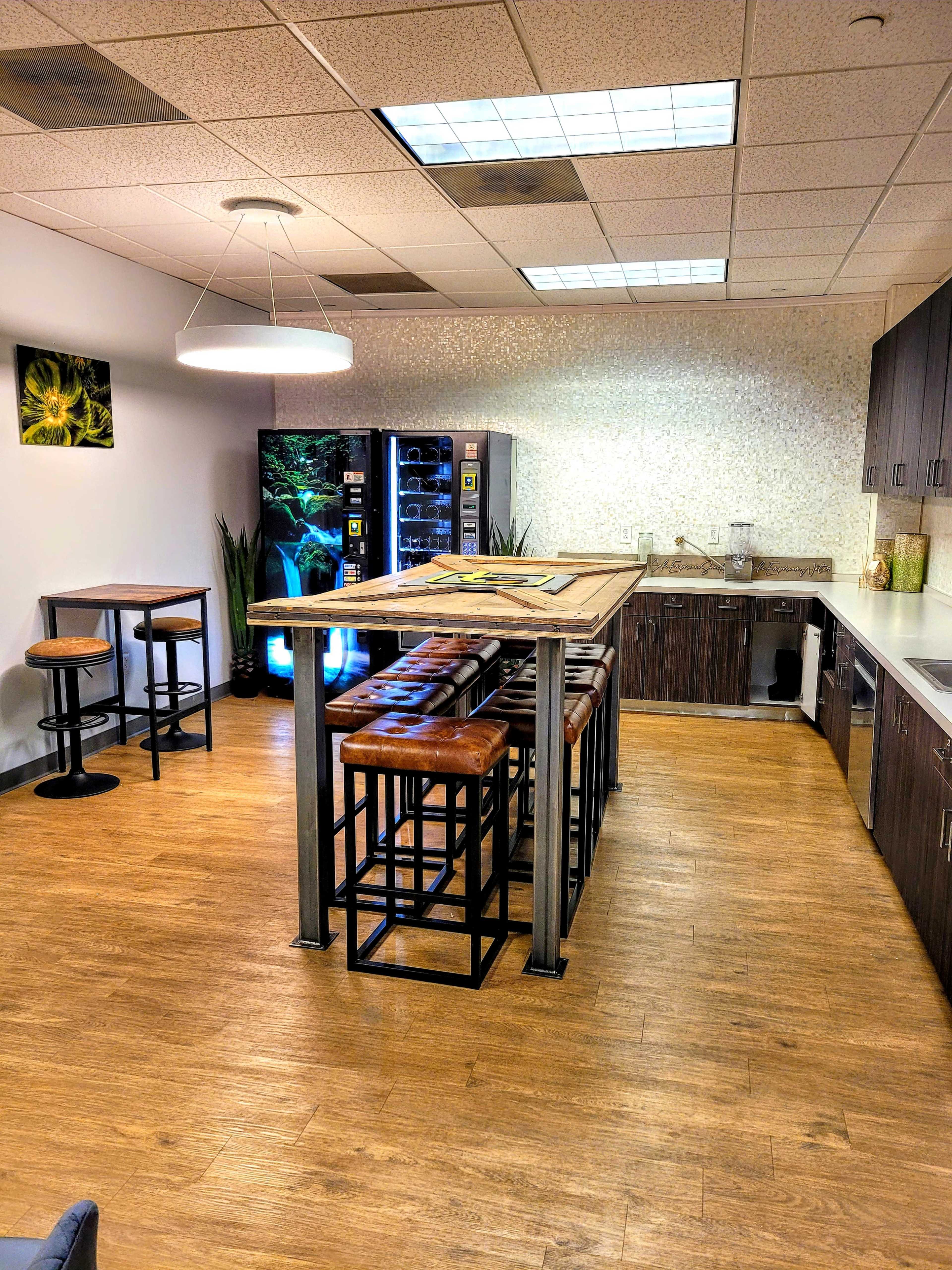 A break room with a large wooden table surrounded by high stools, a vending machine on one side, and a kitchenette area with cabinets.
