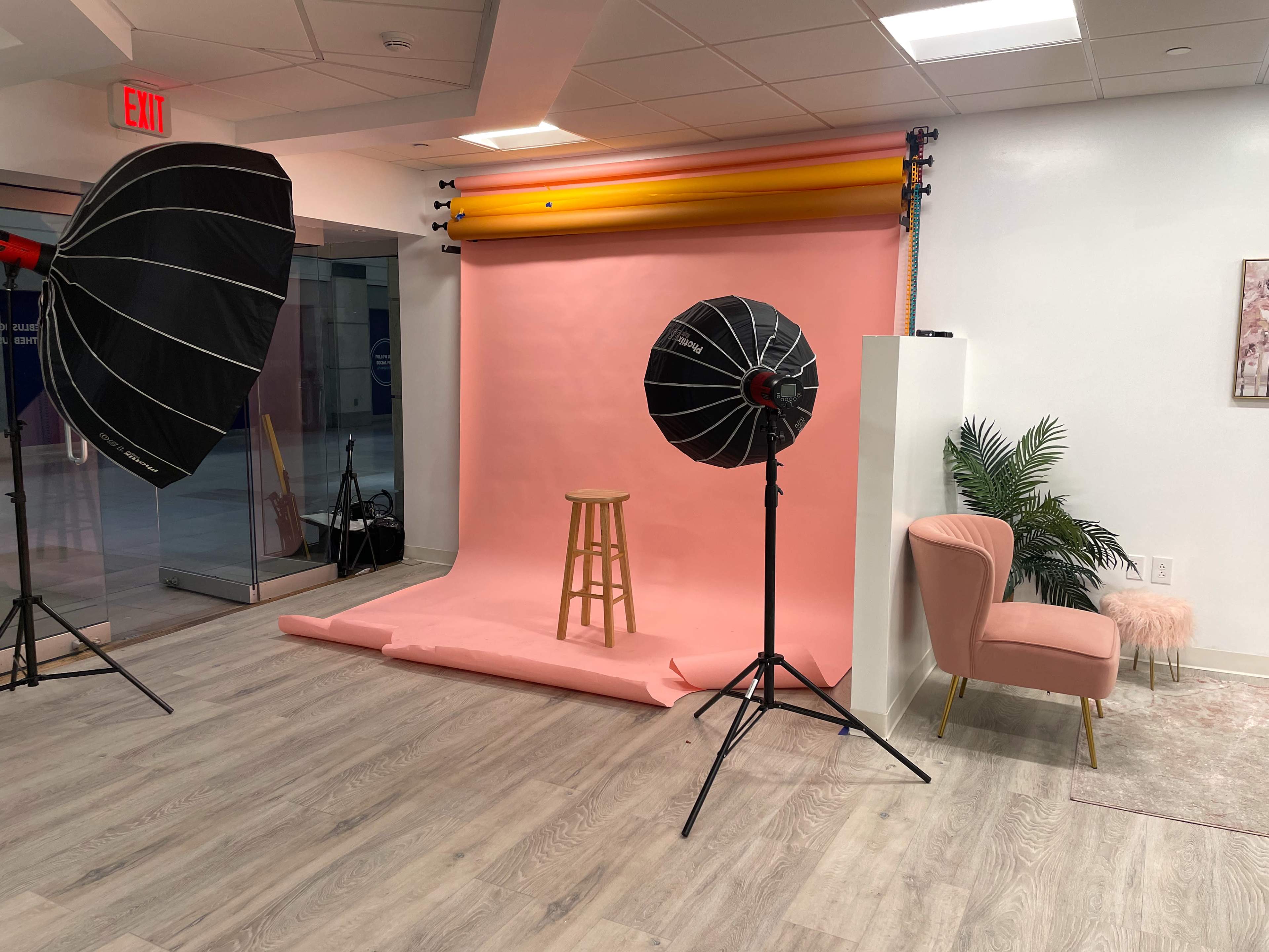 A photography studio setup featuring a pink backdrop, a wooden stool, two softbox lights, and a pink chair with a decorative plant nearby.