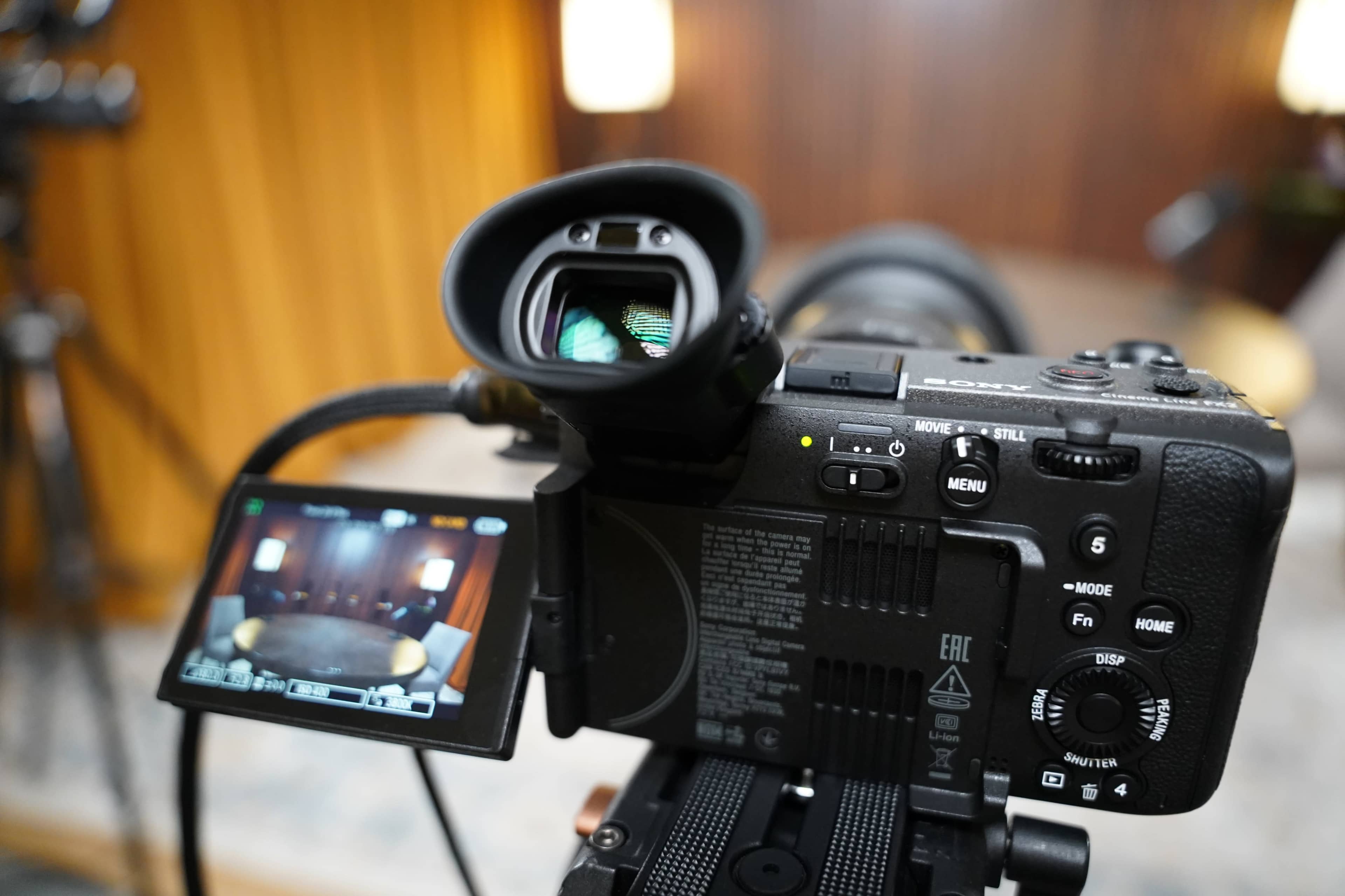 A camera is positioned on a tripod with its display screen and viewfinder visible, set against a blurred background of a room with a wooden wall.