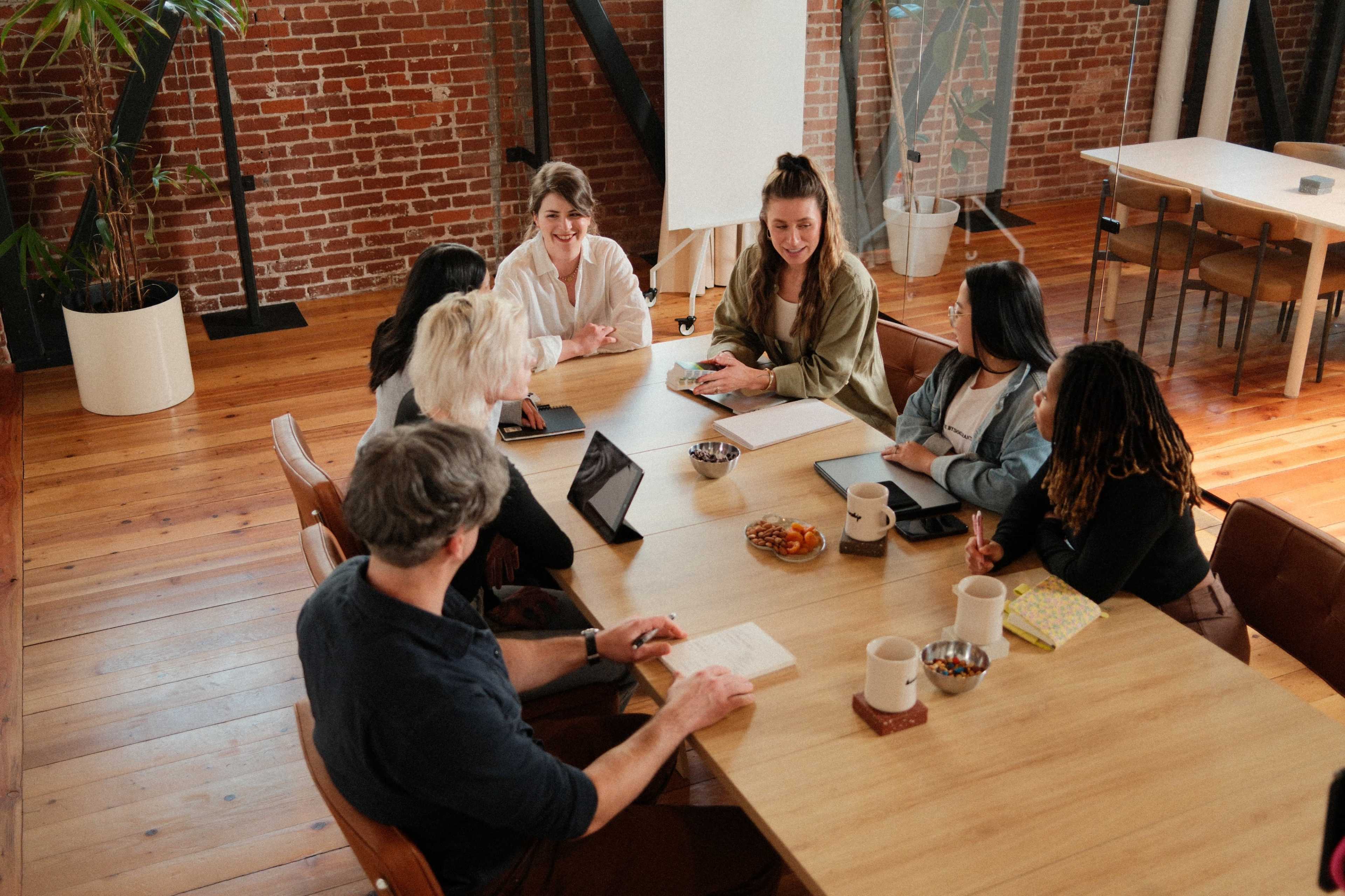 A group of seven people is engaged in a discussion around a wooden conference table in a spacious, well-lit room with exposed brick walls.