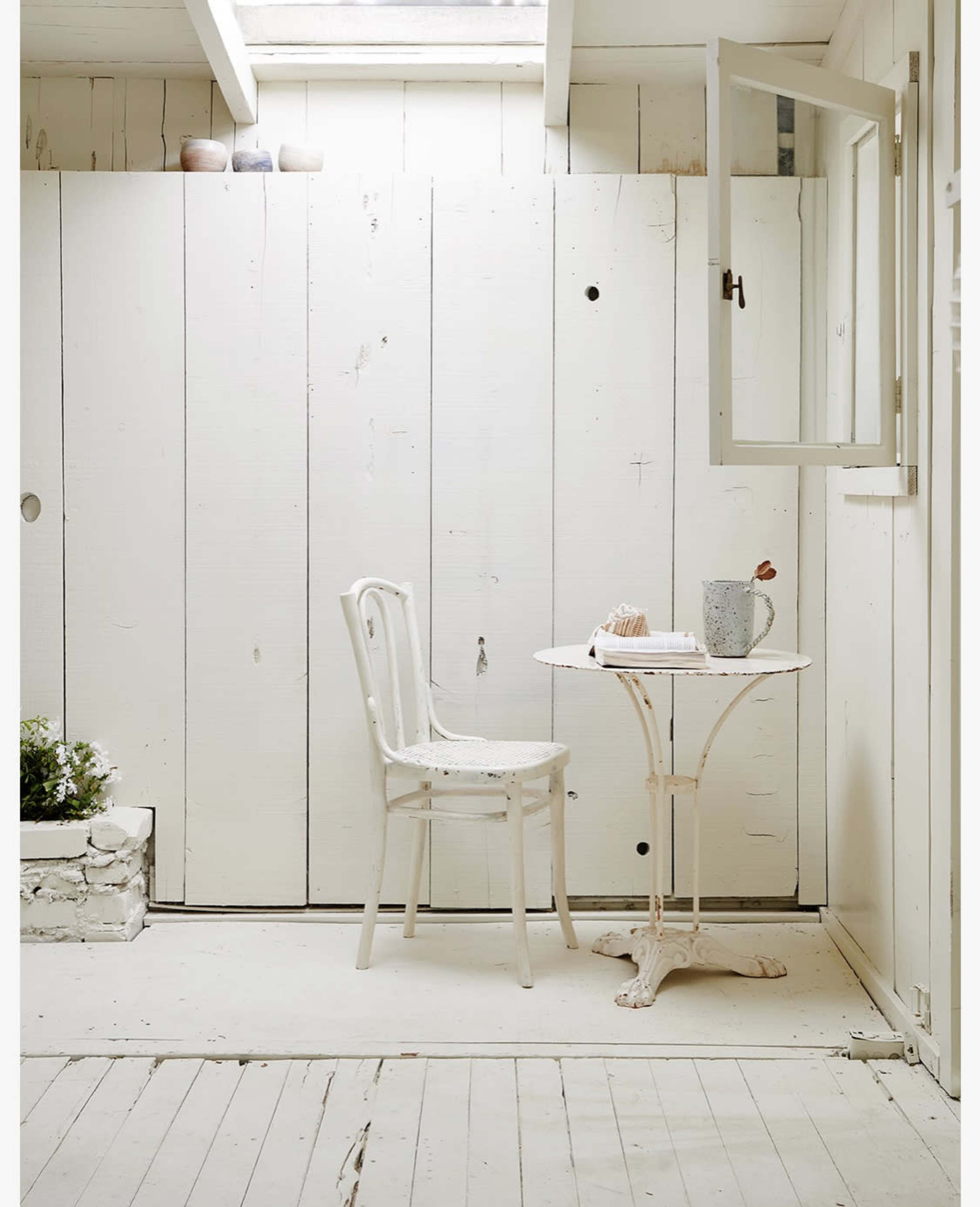 A small, white room features a round table with a chair and a mug, beside a window and a potted plant.