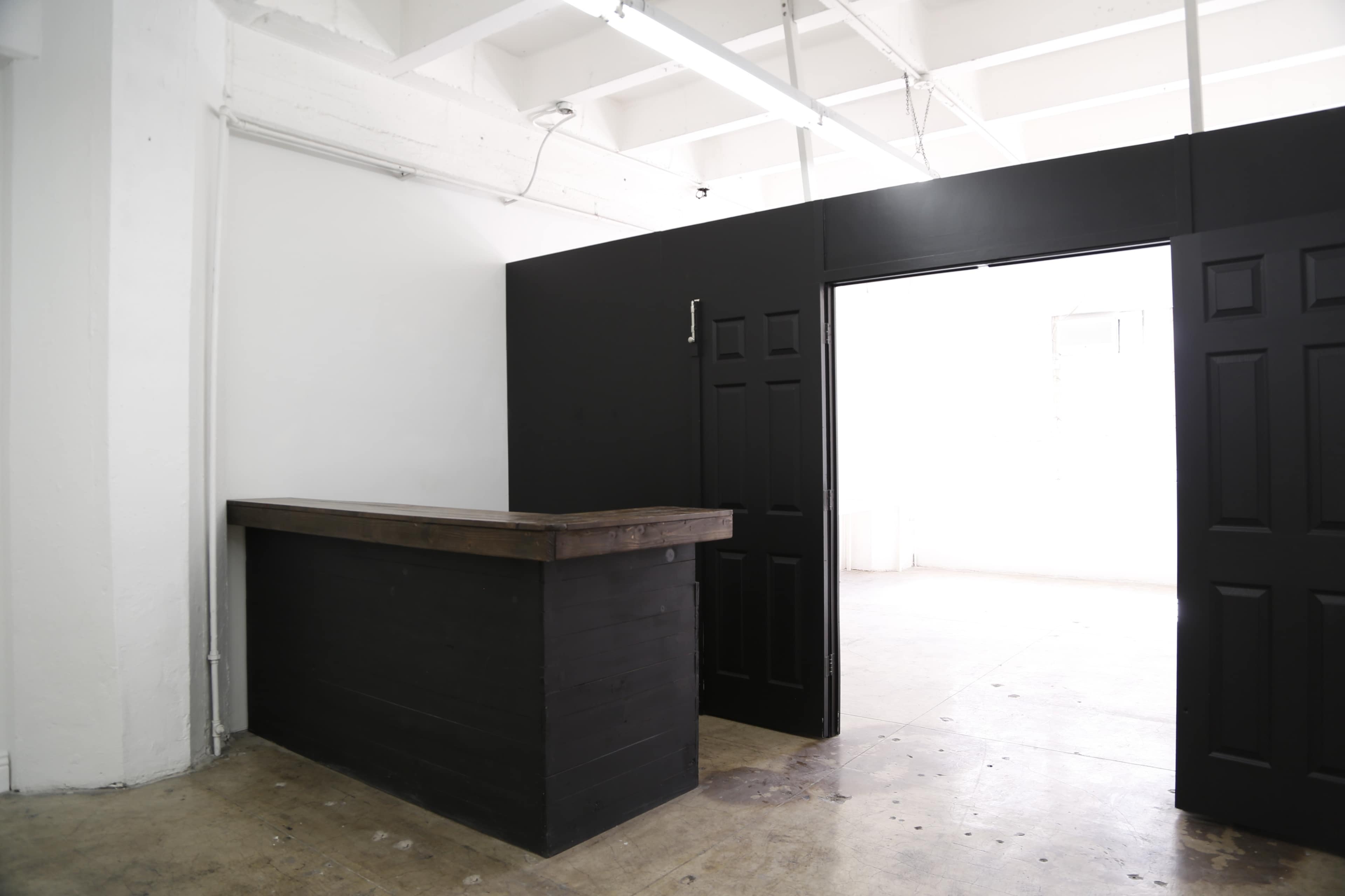 A minimalist reception area with a wooden counter against a black wall and an open doorway leading to a bright space.