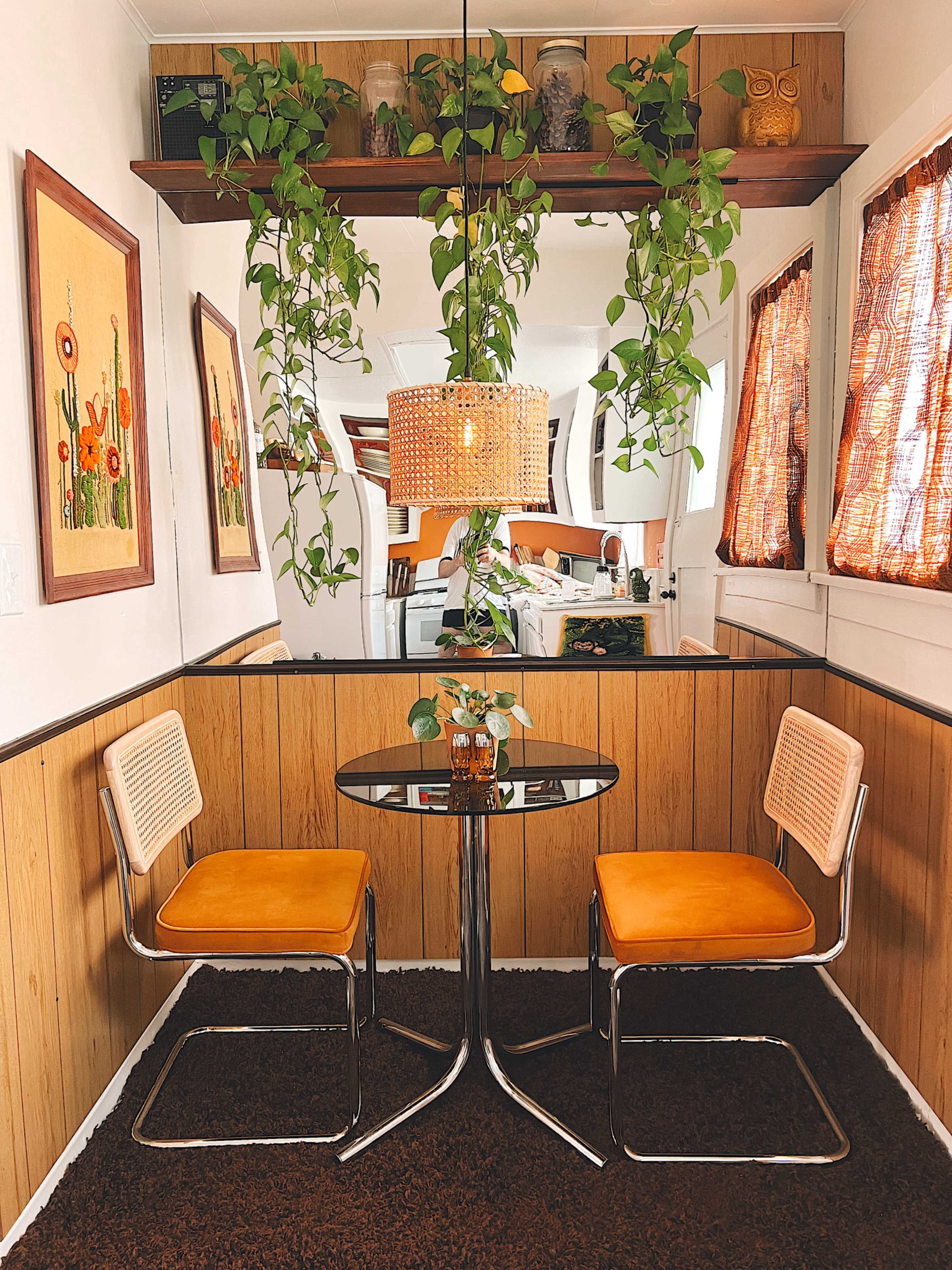 A small dining area features two orange chairs at a glass table, with hanging plants and framed floral artwork on the walls.