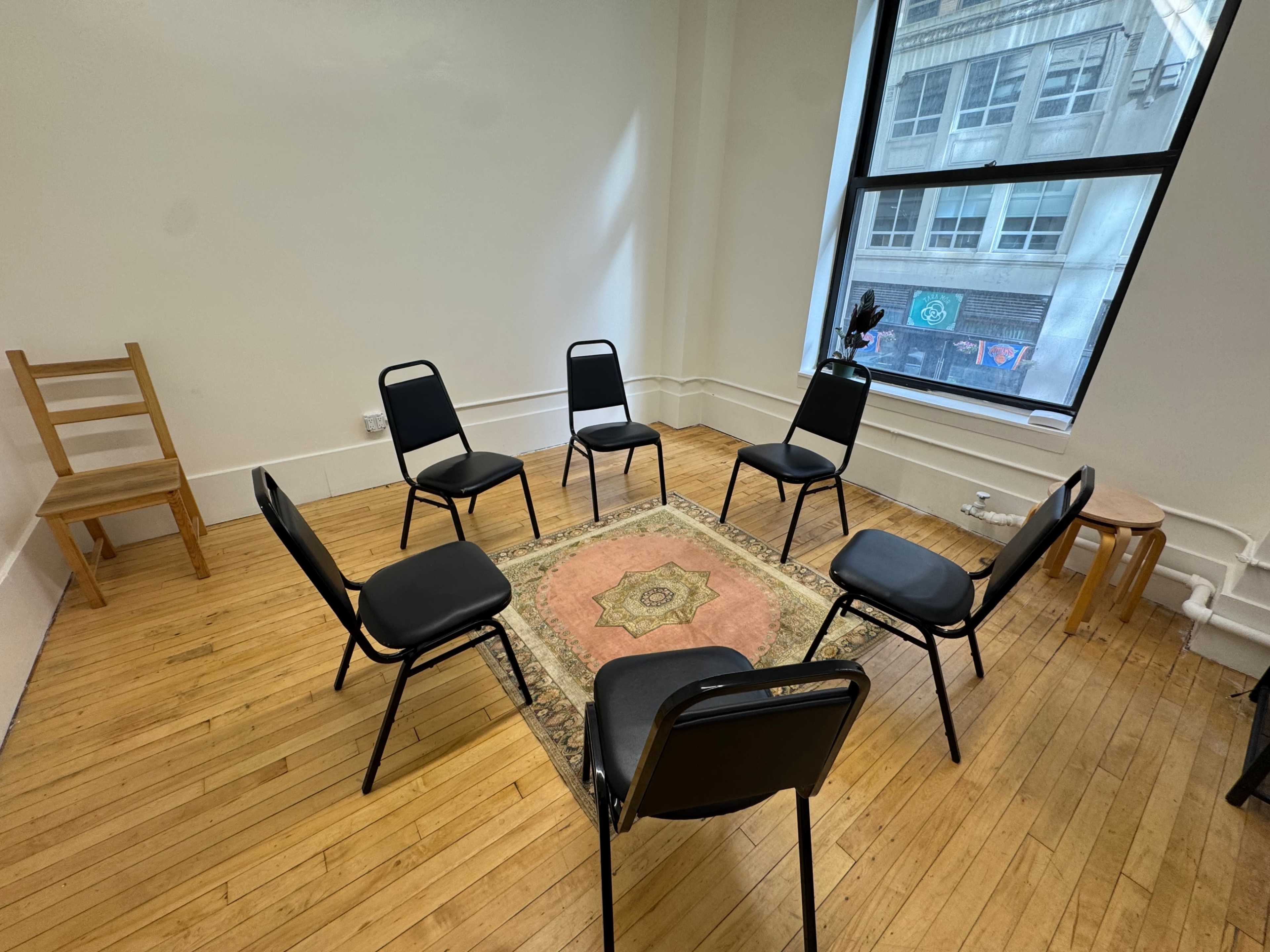 The image shows a group of six black chairs arranged in a circle around a patterned rug on a wooden floor in a well-lit room with a large window.