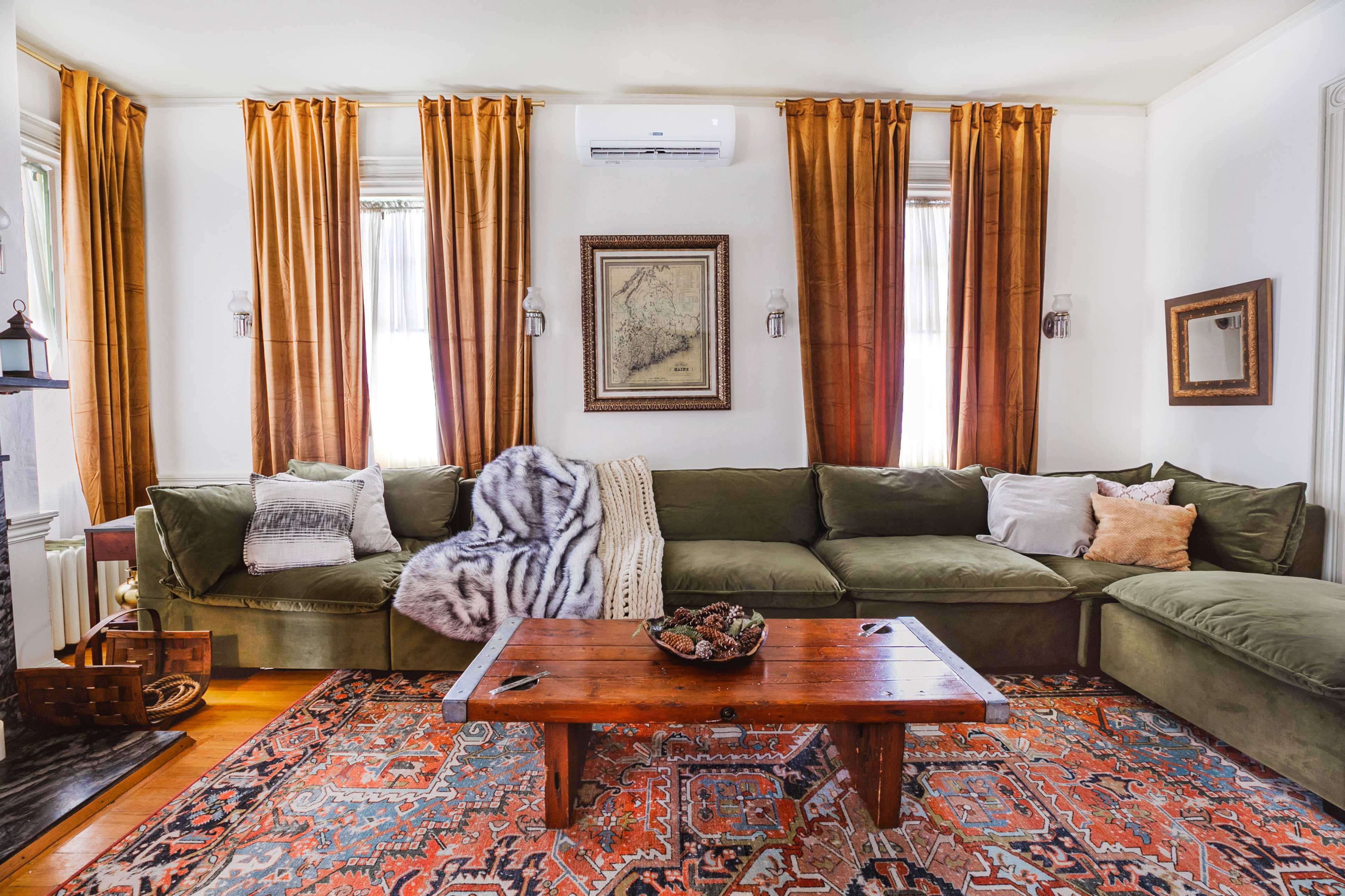 The image shows a living room with a large green sectional sofa, a wooden coffee table, and orange curtains hanging by the windows.