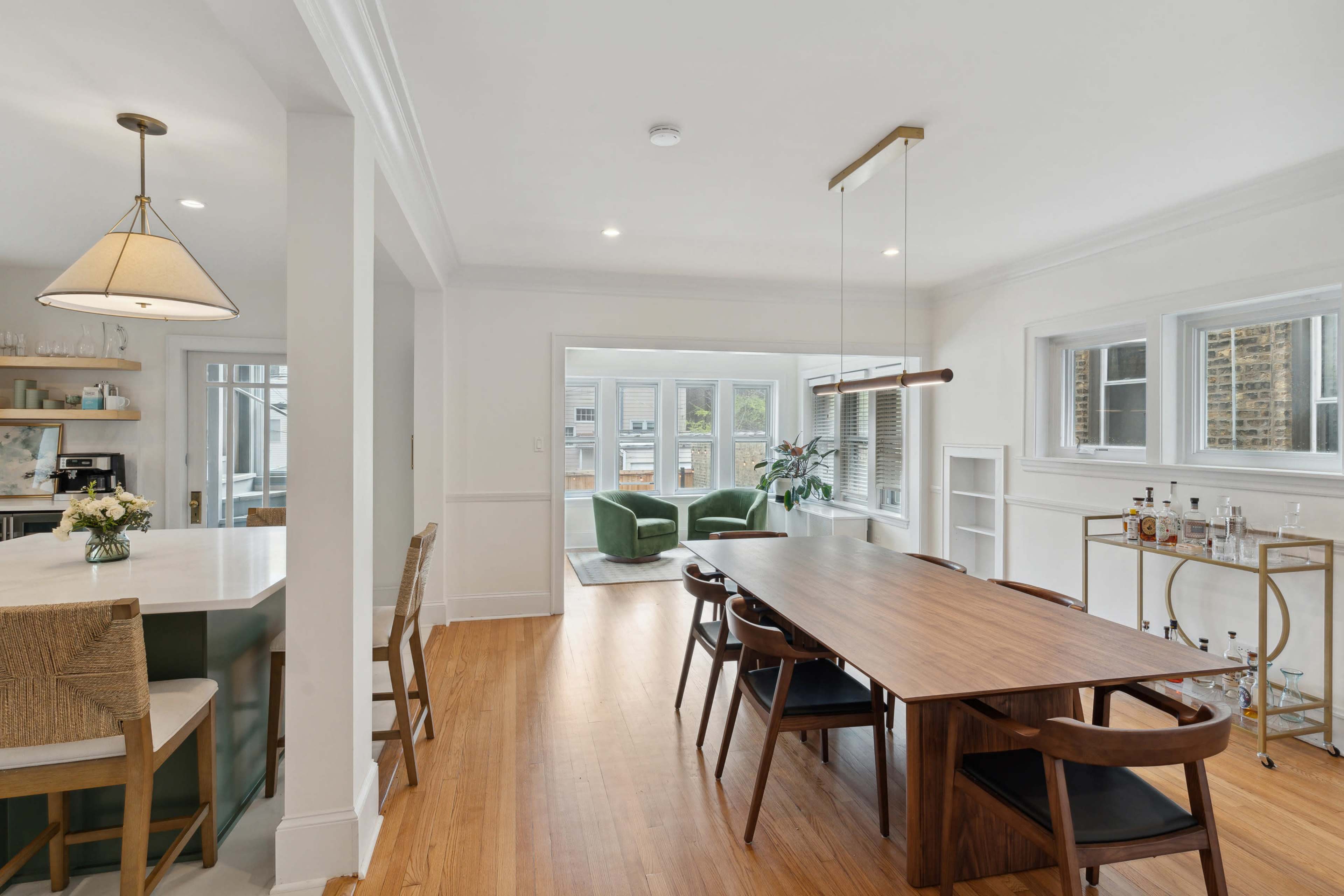 The image shows a spacious modern dining area with a long wooden table, surrounded by chairs, and a view of a well-lit living space with green chairs and large windows.