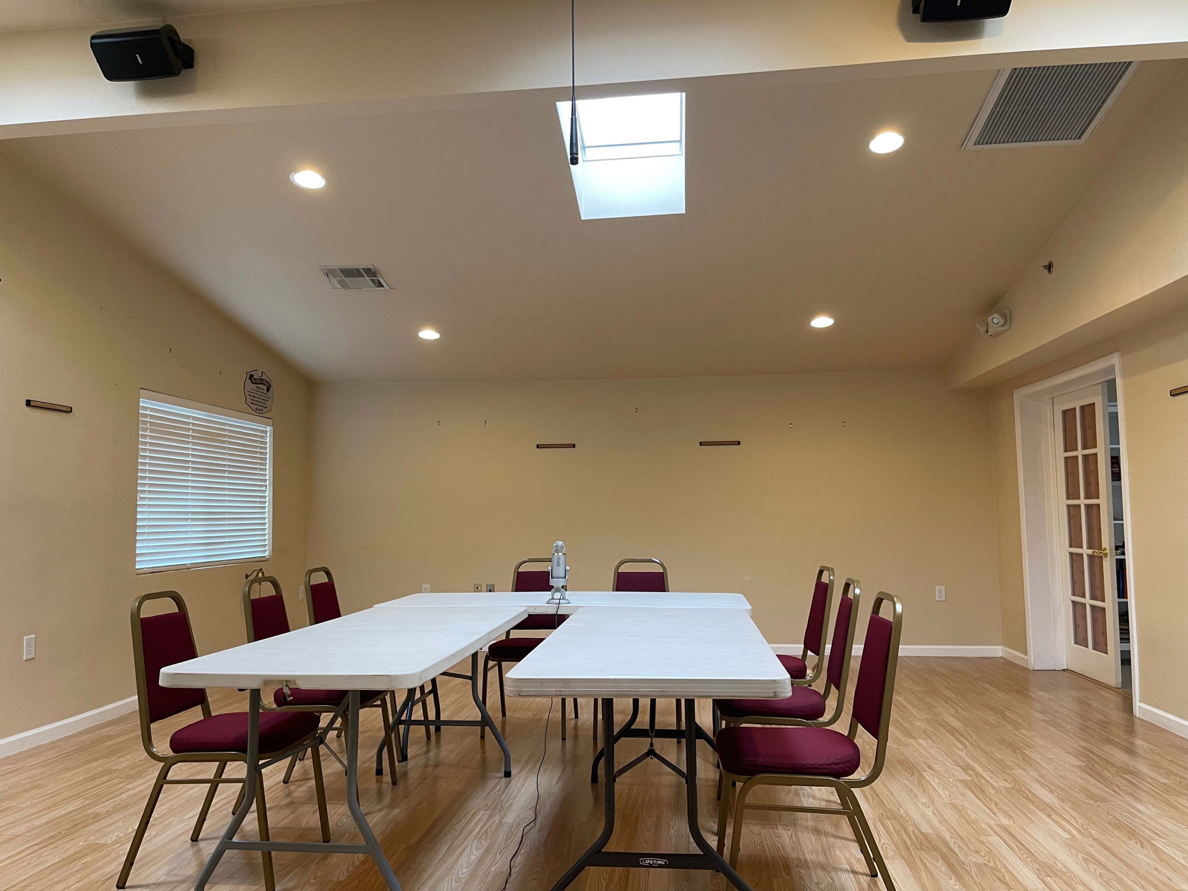 A meeting room features two rectangular tables arranged in a U-shape, surrounded by chairs, with a skylight and windows letting in natural light.
