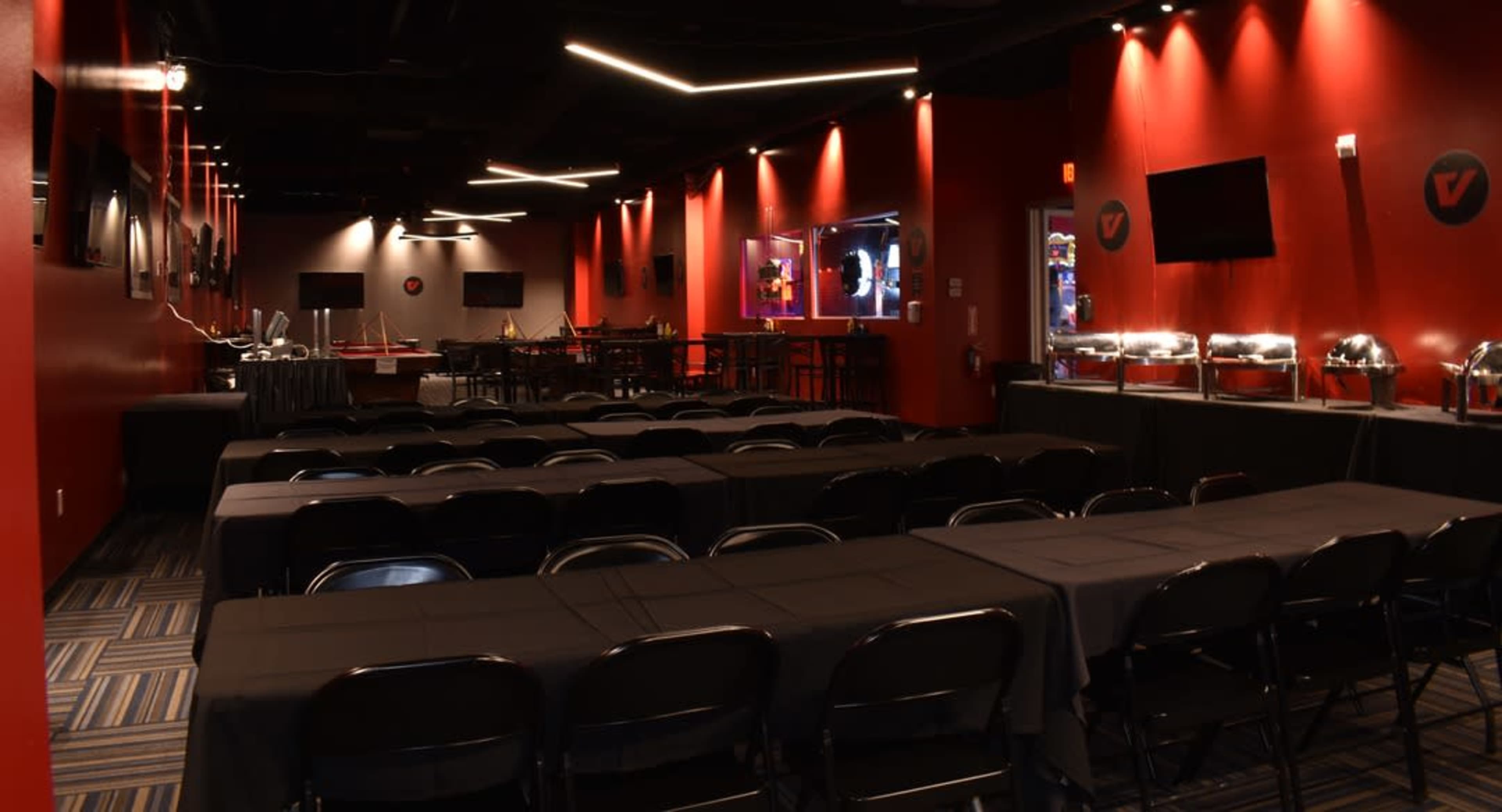 The image depicts a spacious red-accented event room with black tables arranged in rows, equipped with multiple screens and a buffet area along one wall.