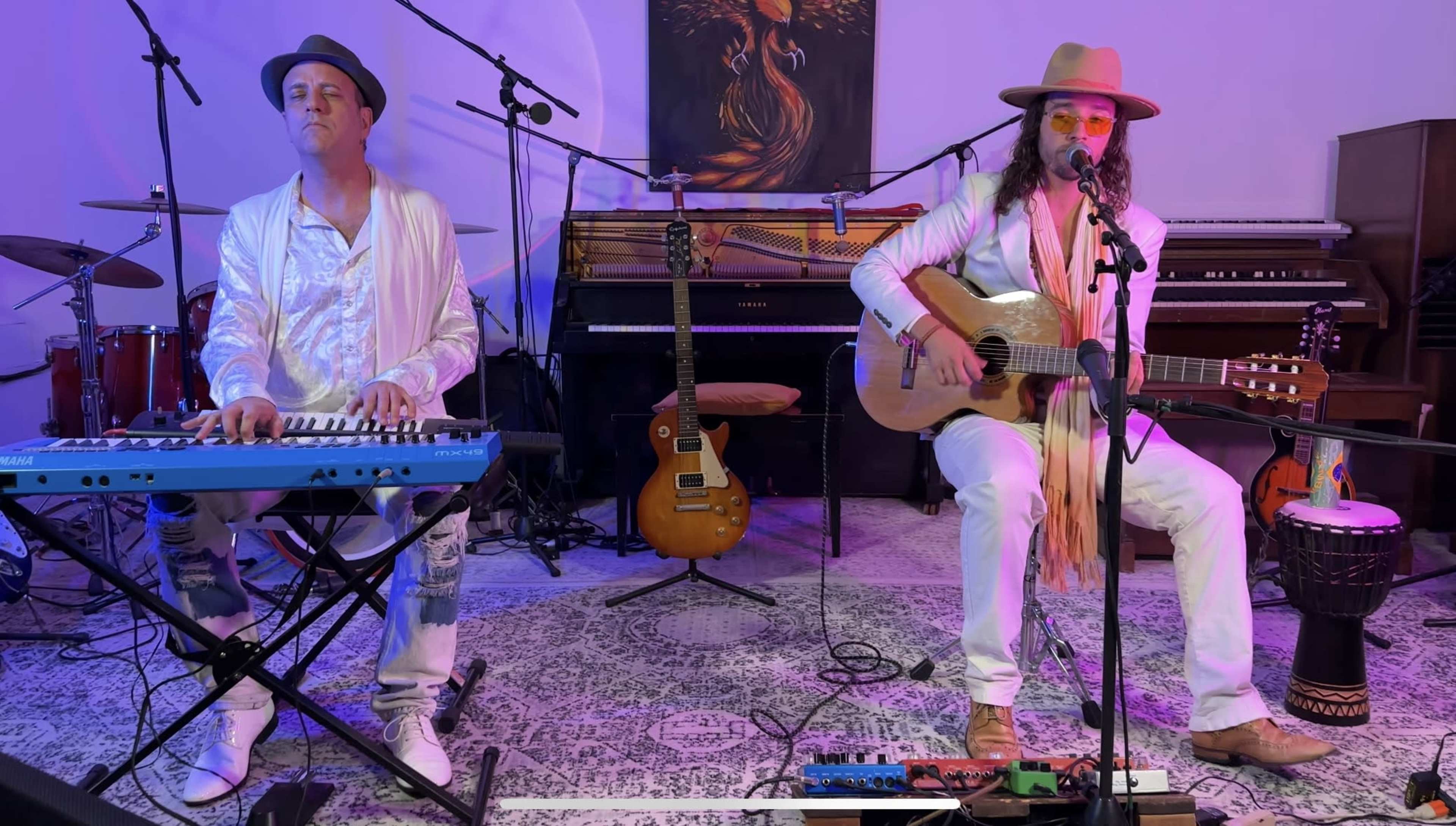 Two musicians perform on stage, one playing a keyboard and the other strumming an acoustic guitar, with a piano and guitars in the background.