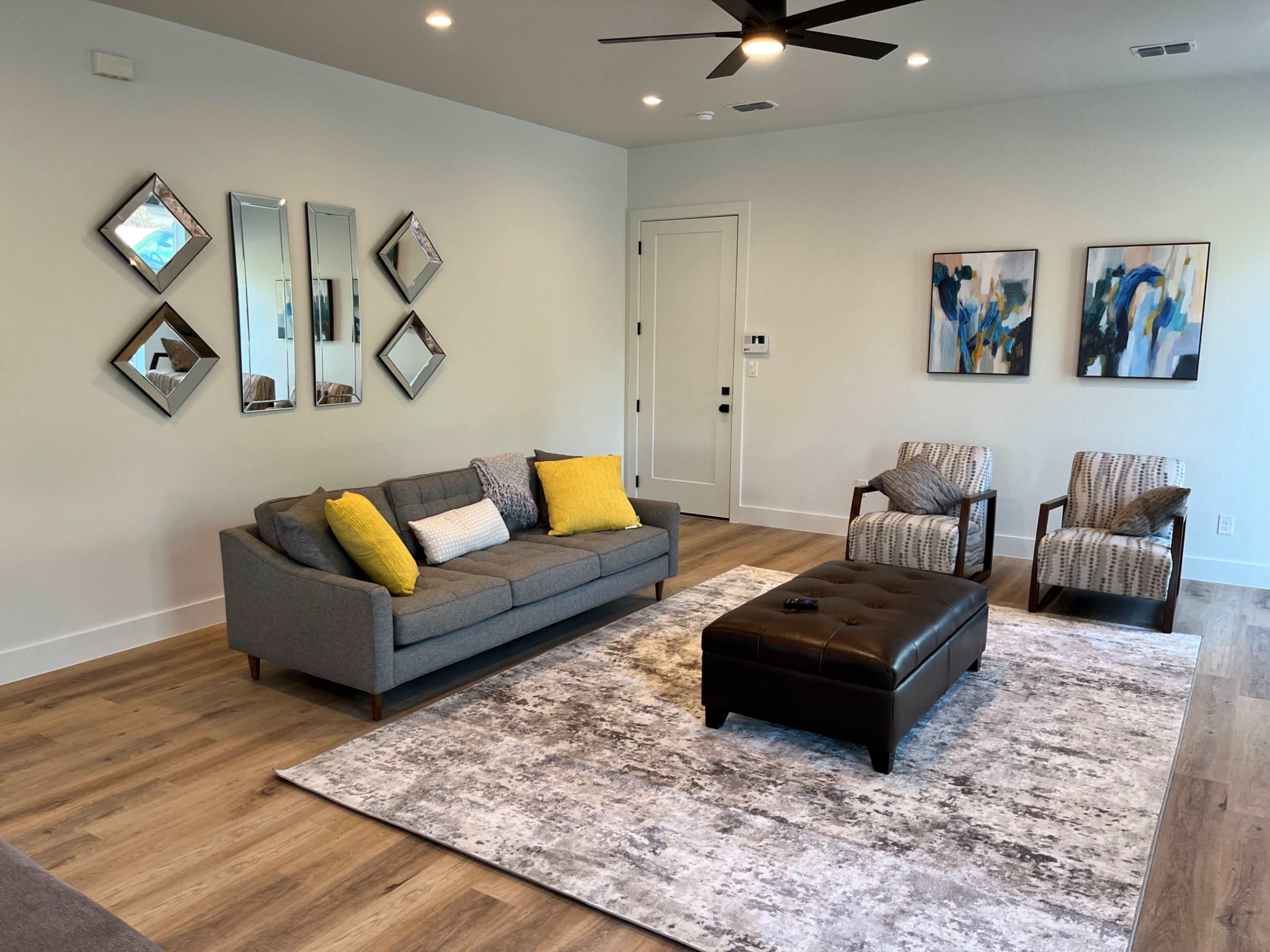 A modern living room featuring a gray sofa with yellow cushions, two patterned armchairs, a large area rug, and three decorative mirrors on the wall.