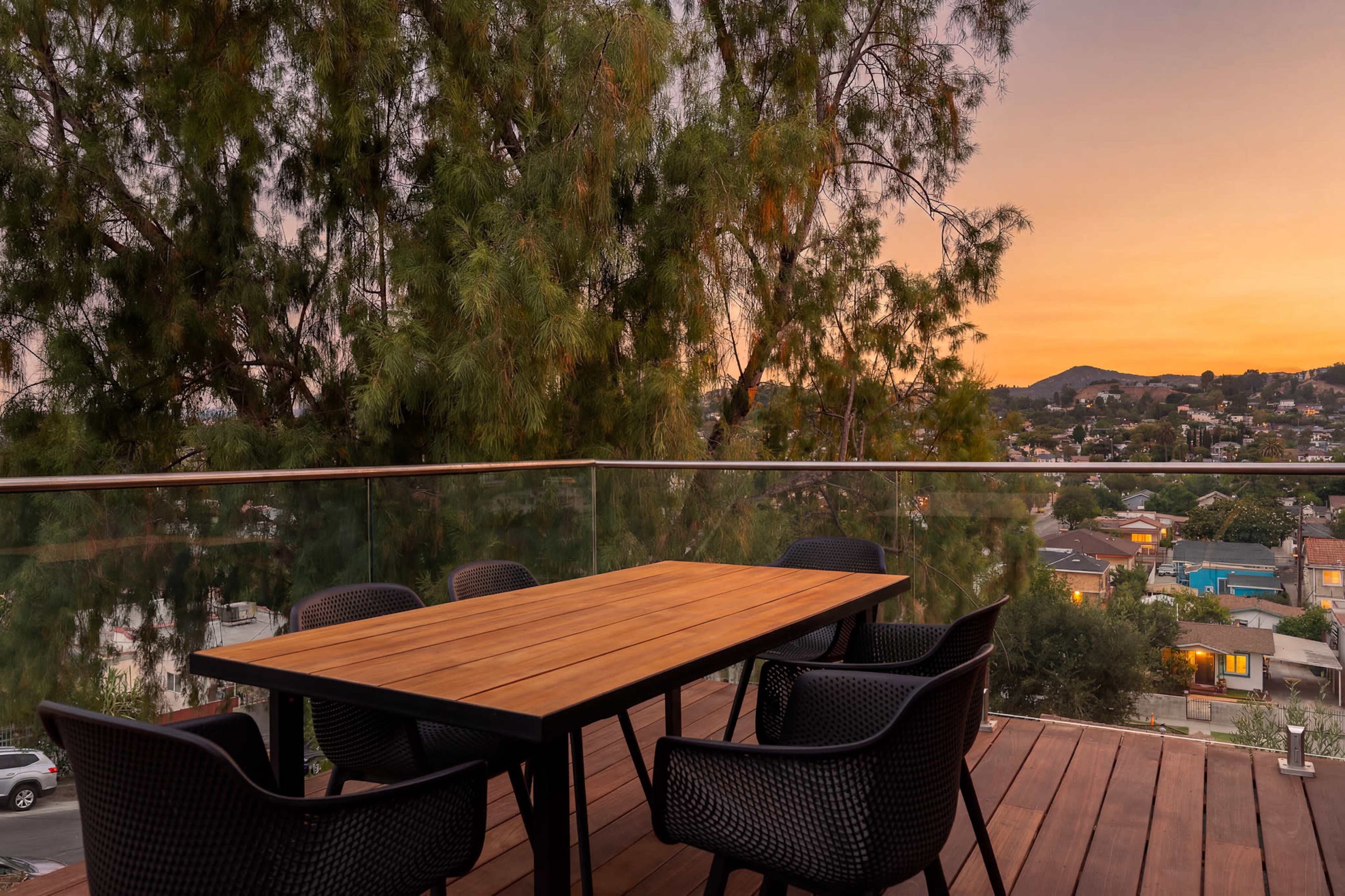 A wooden dining table is set on a balcony surrounded by black chairs, with a view of a neighborhood at sunset.
