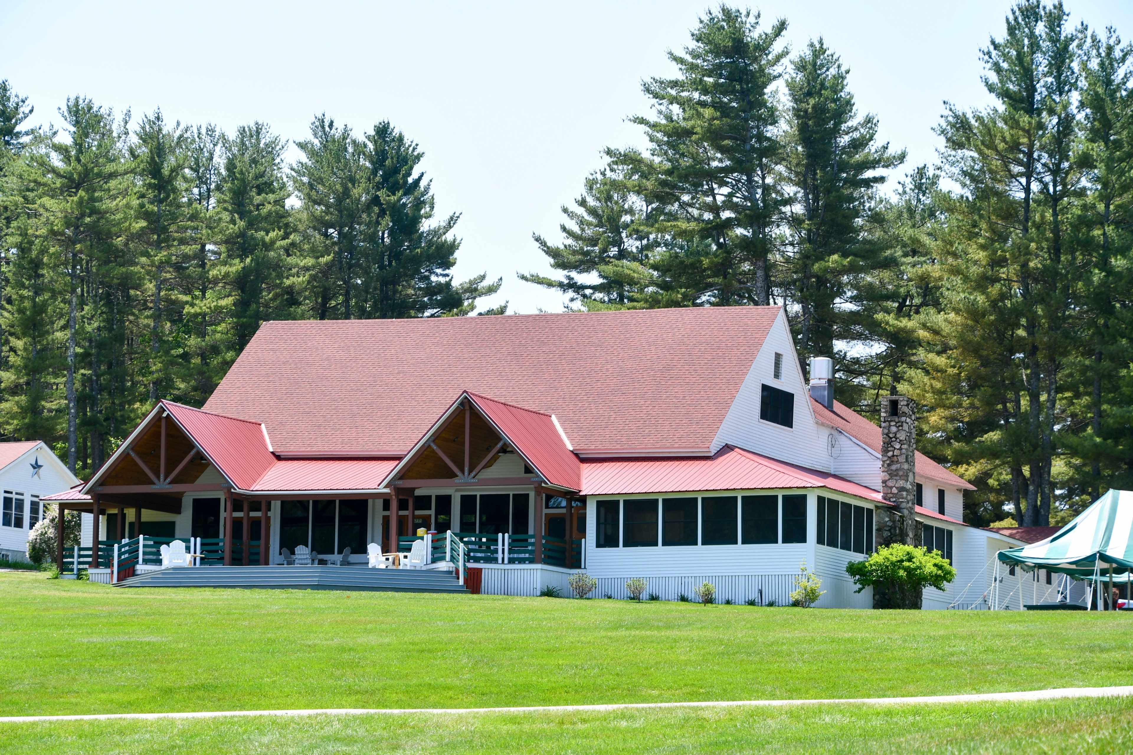 A large white house with a red roof and multiple porches is set against a backdrop of tall pine trees.