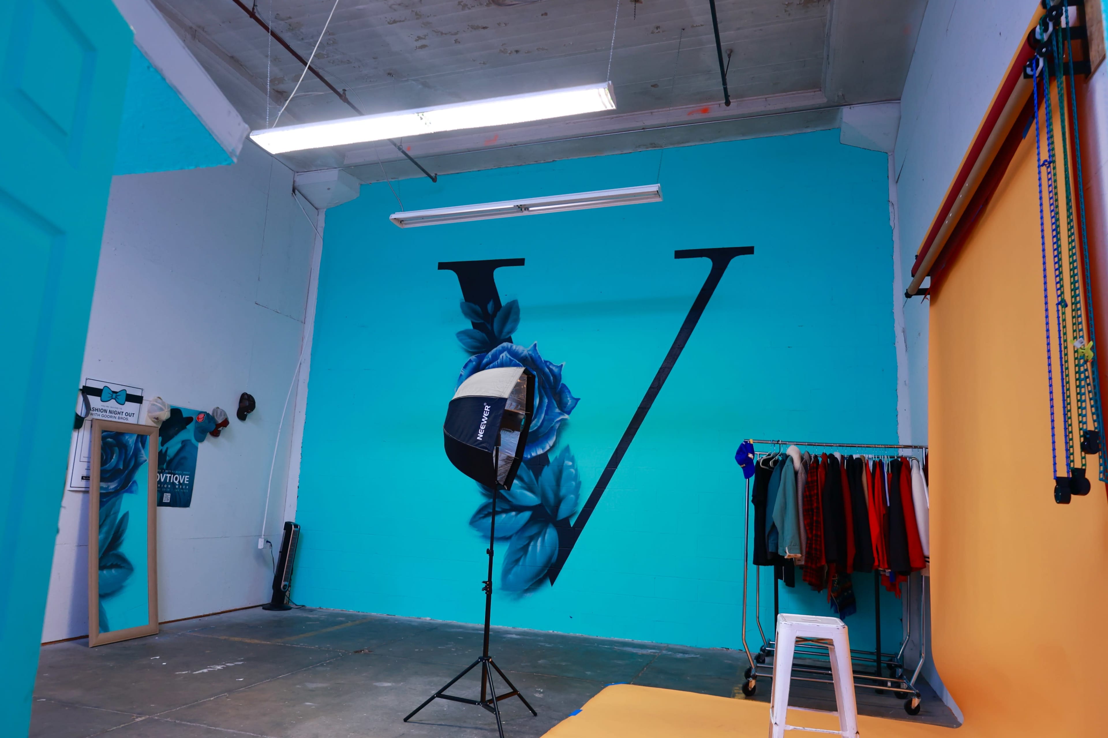 A photography studio with a bright blue wall featuring a large letter "V" and a rack of clothing alongside a lighting setup.