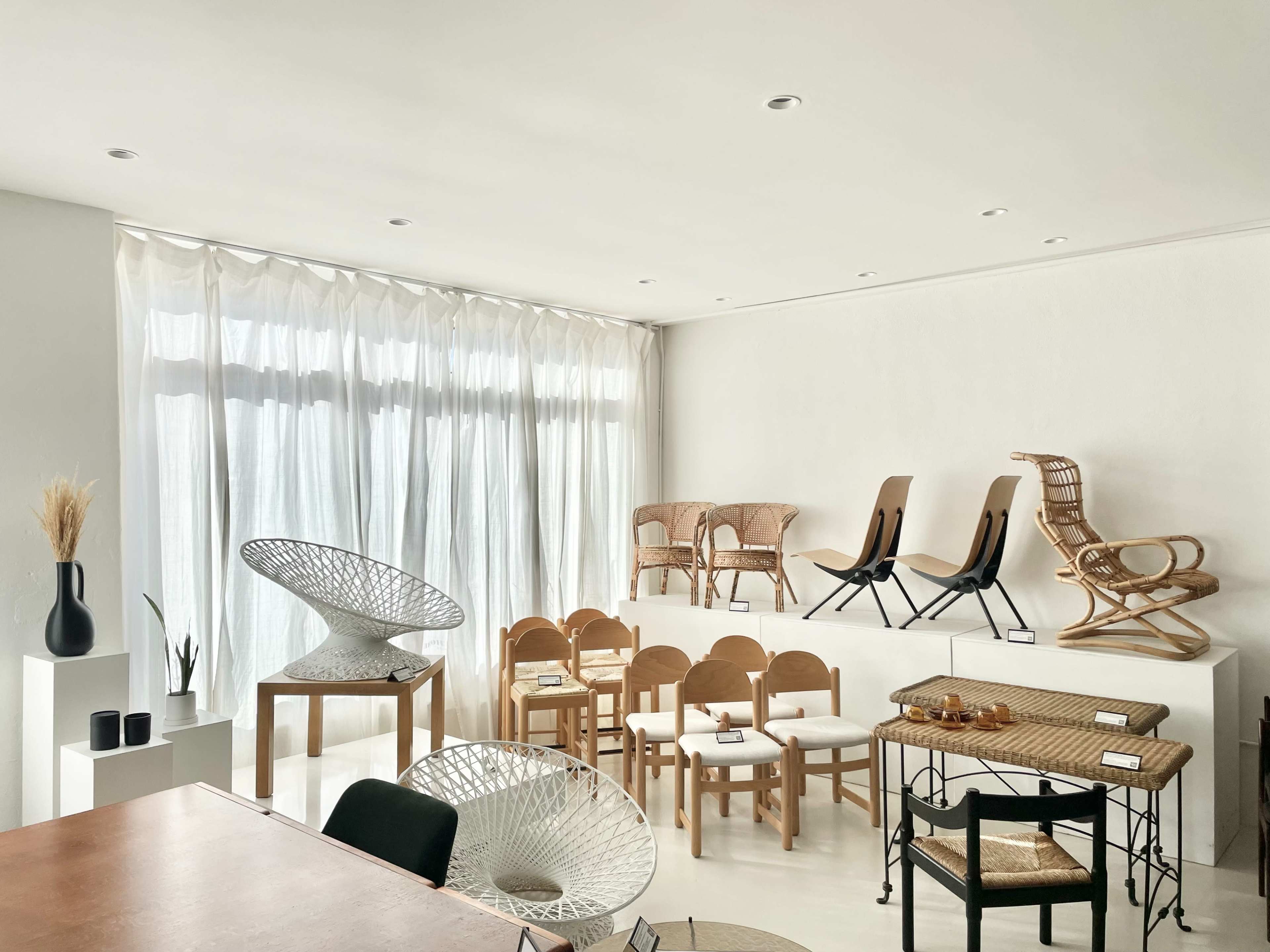 A modern, minimalist interior space featuring various wooden and wicker chairs, a round sculpture, and large windows with sheer curtains.