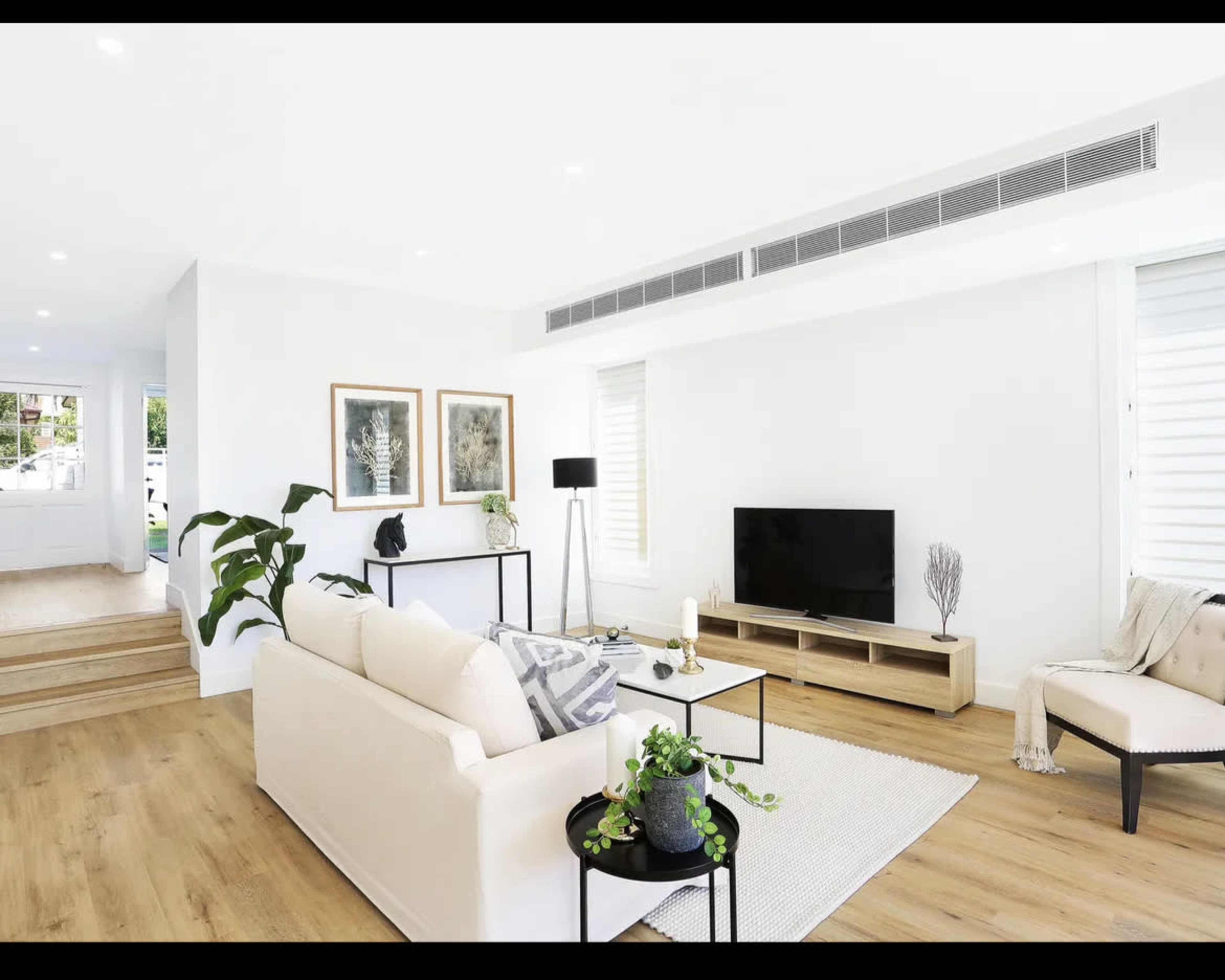 A bright living room featuring a cream-colored sofa, a black coffee table, a television stand, and large windows with white shutters.