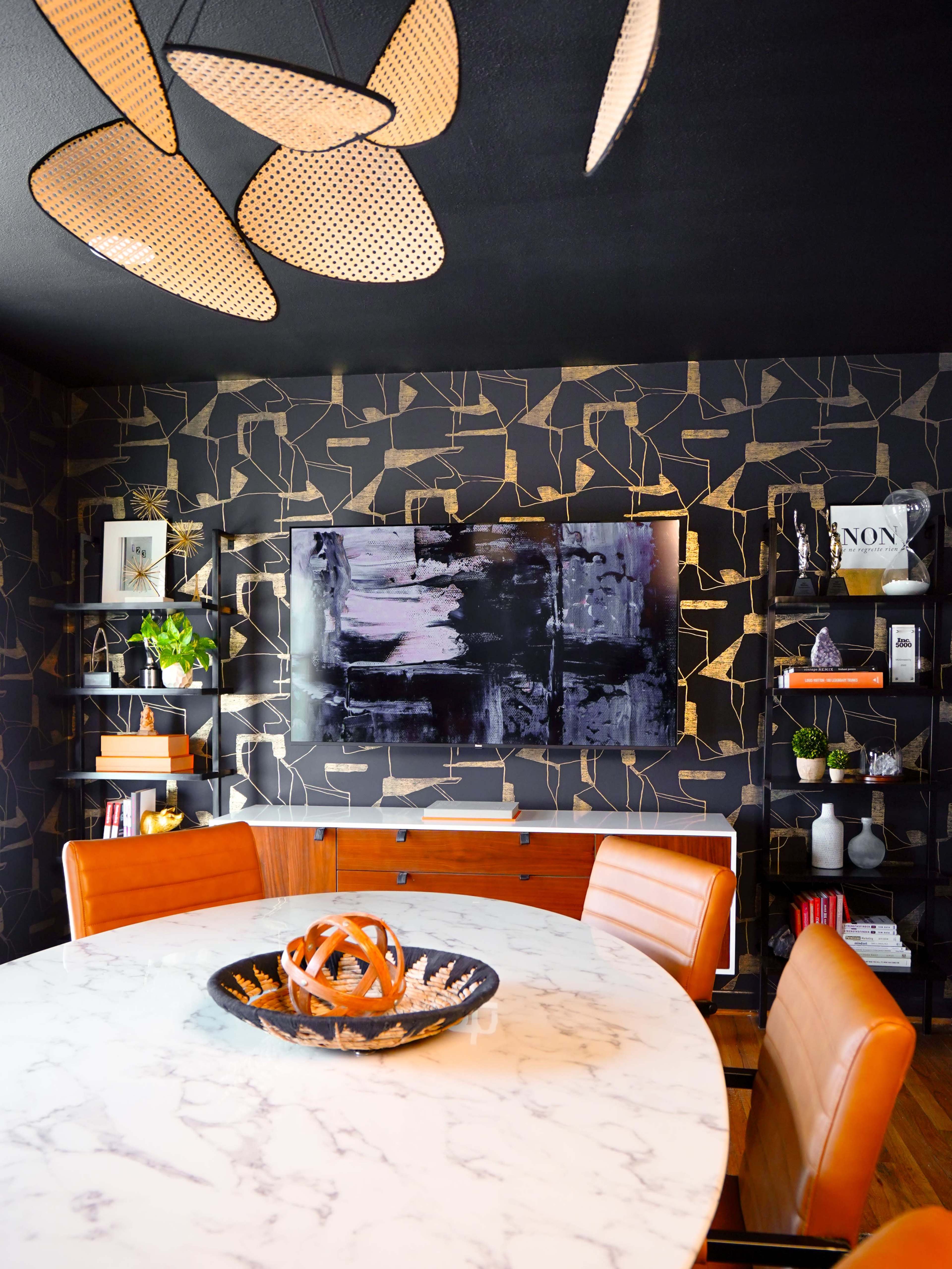 The image showcases a modern dining area with a round marble table, wooden chairs, and a dark geometric-patterned wall featuring a flat-screen TV and decorative shelves.