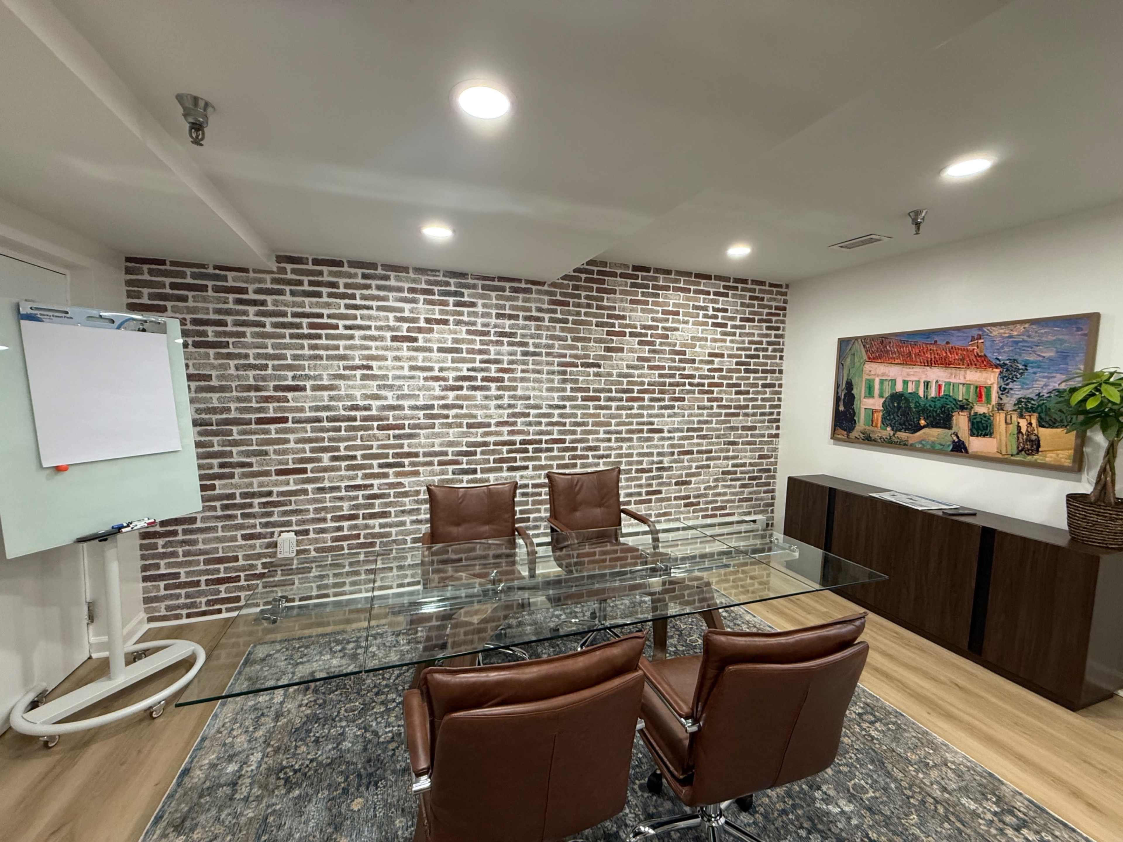 The image shows a modern conference room with a glass table, brown leather chairs, a whiteboard on a stand, and a brick accent wall.