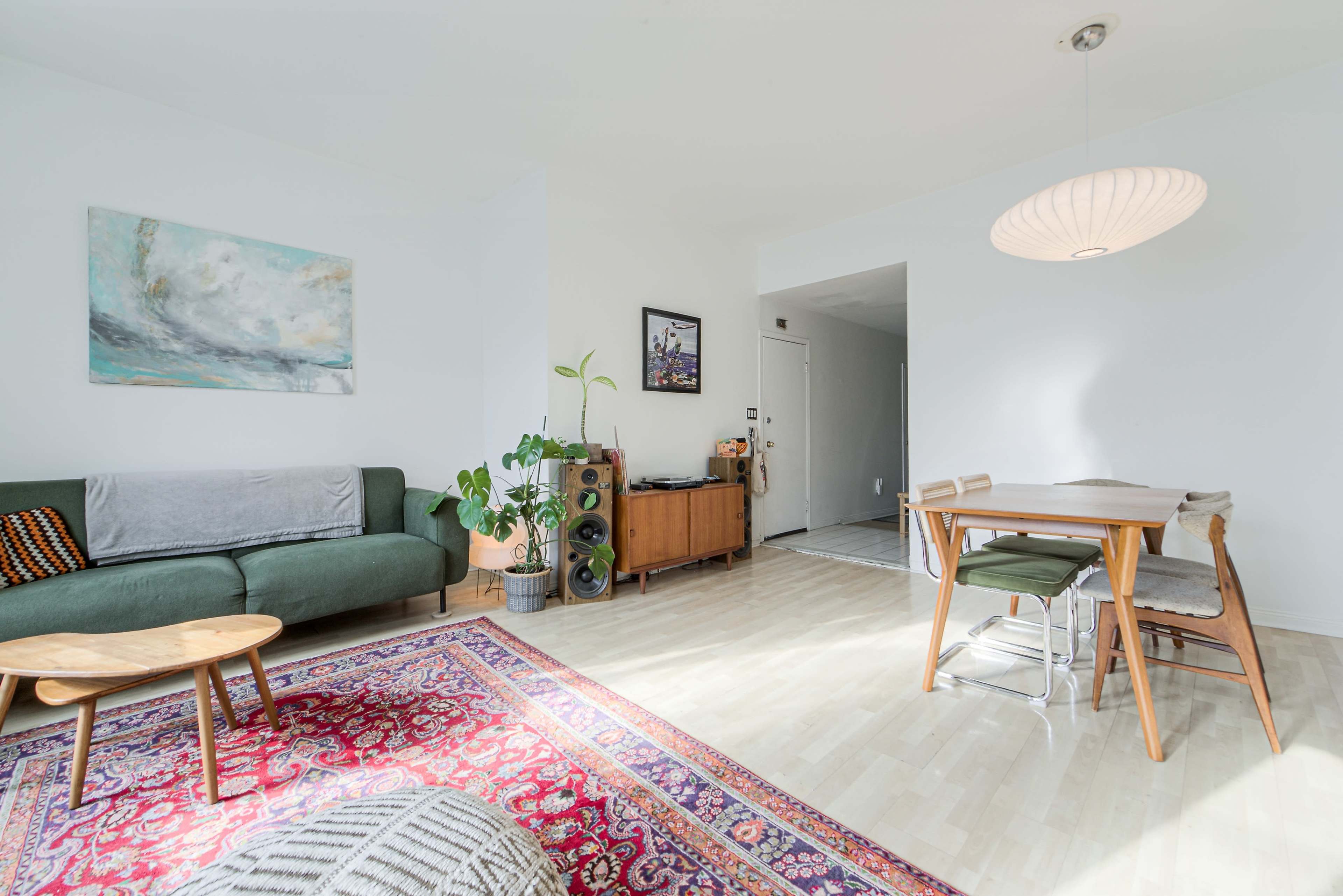 The image shows a bright living space with a green sofa, a wooden dining table, and a colorful area rug.