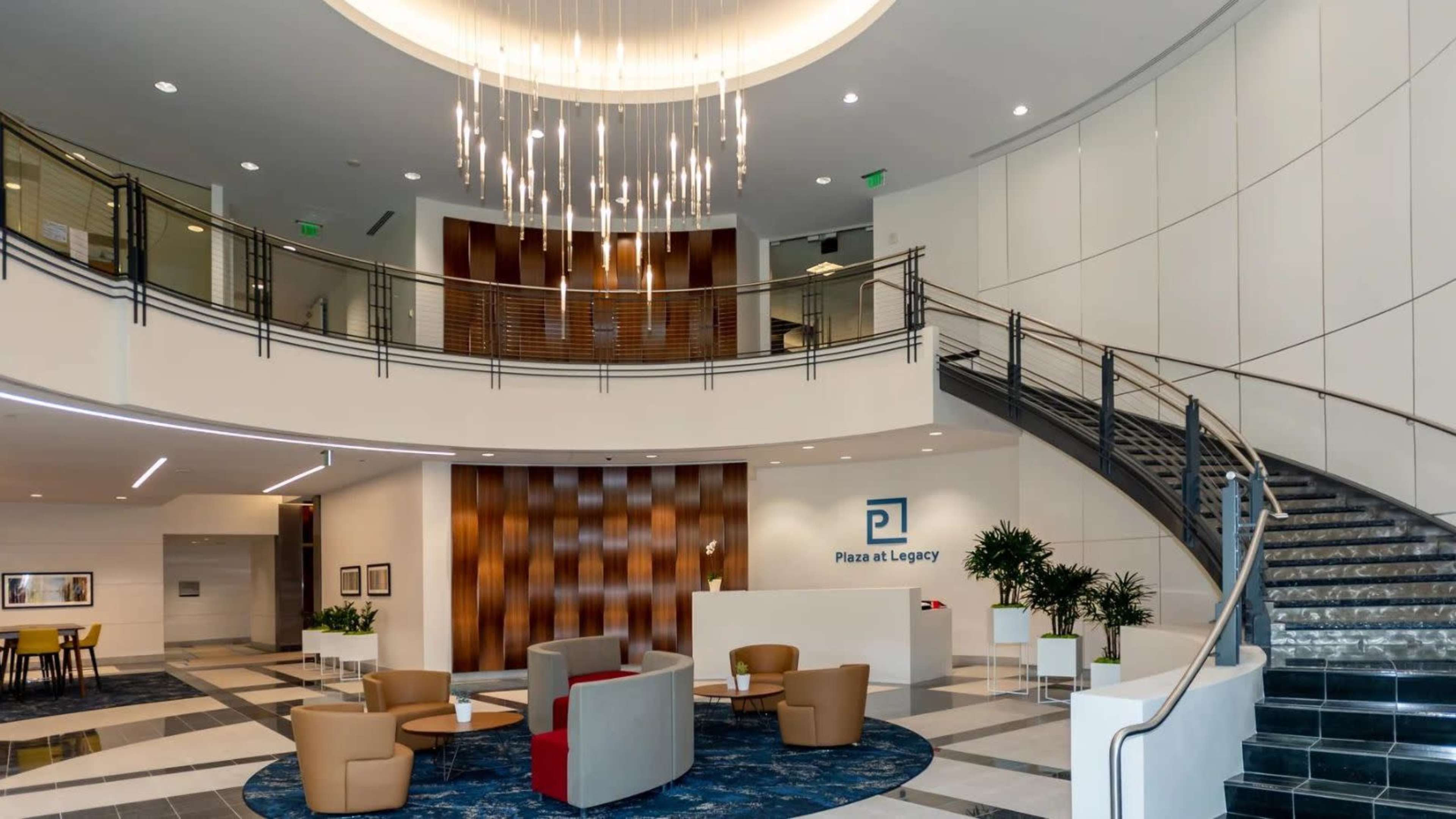 The image shows the interior of a modern office lobby featuring a spiral staircase, seating area with chairs, and a reception desk with the name "Plaza at Legacy" prominently displayed.