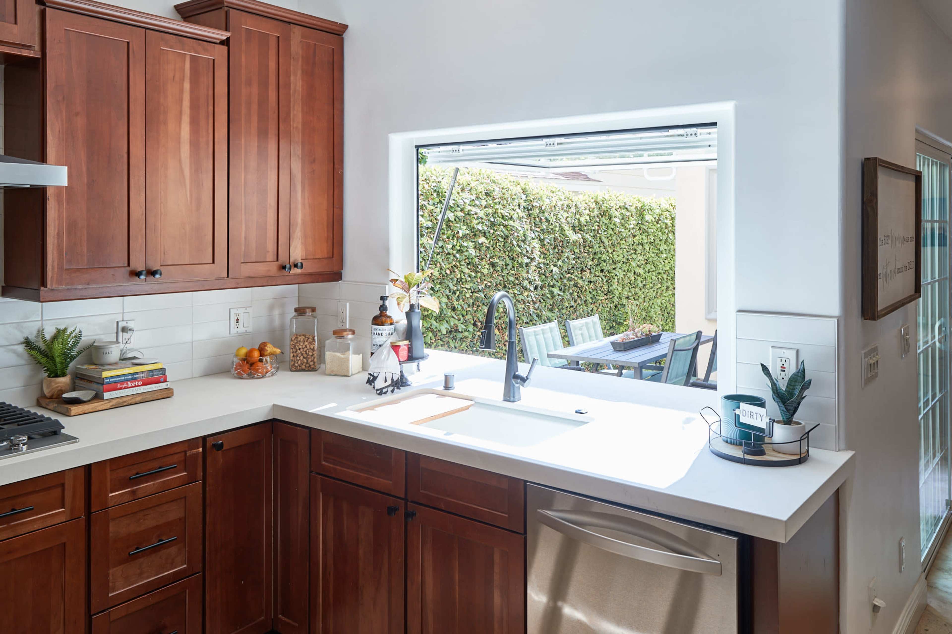 A kitchen with wooden cabinets and a white countertop features a window that opens to a backyard with green foliage.