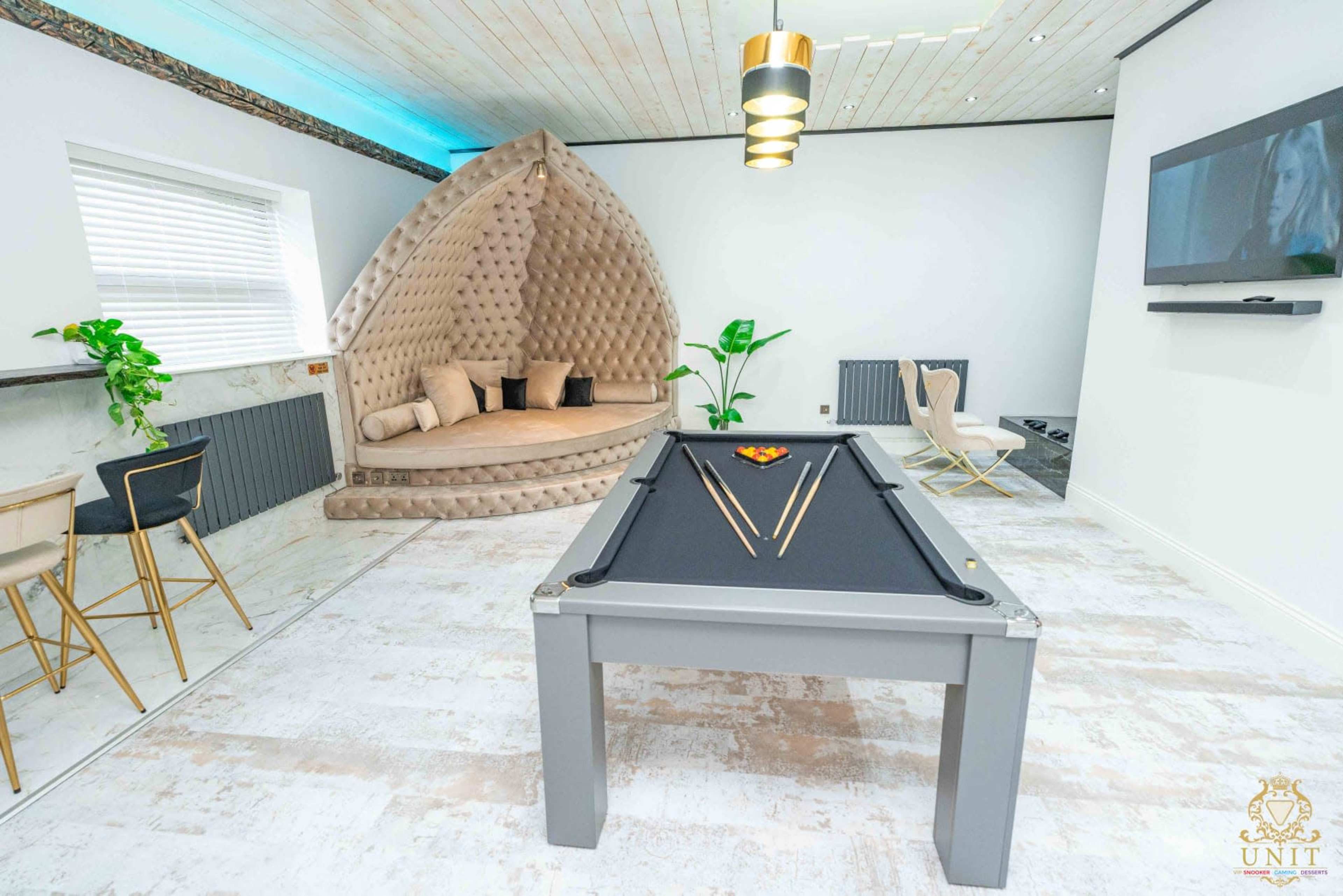 The image shows a modern recreational room featuring a billiard table in the center, surrounded by a curved, cushioned seating area and minimalist decor.