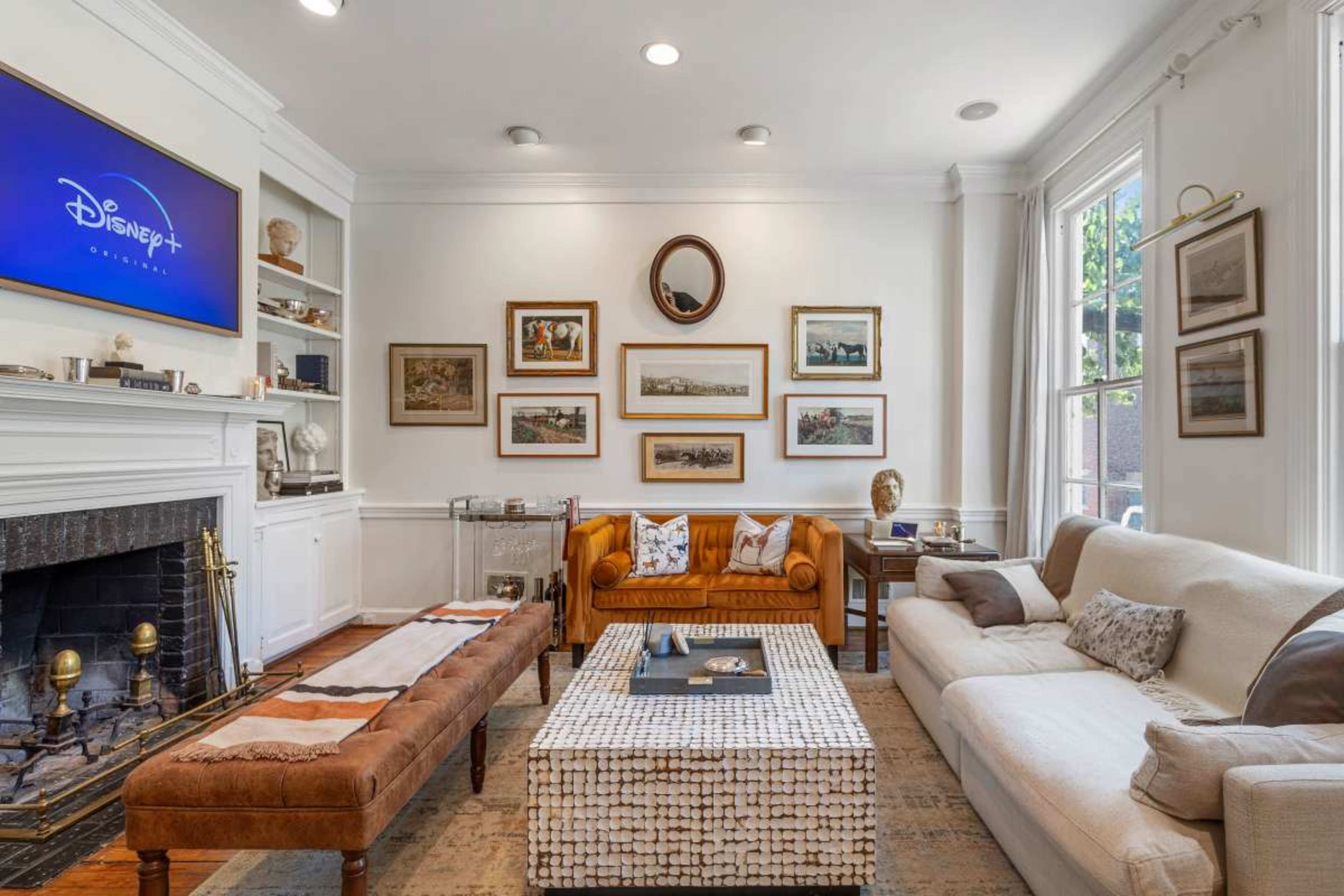 The image shows a cozy living room featuring a mix of seating options, including a tan leather couch, a modern upholstered sofa, and a wooden coffee table, with framed artwork on the walls and a television displaying the Disney+ logo.