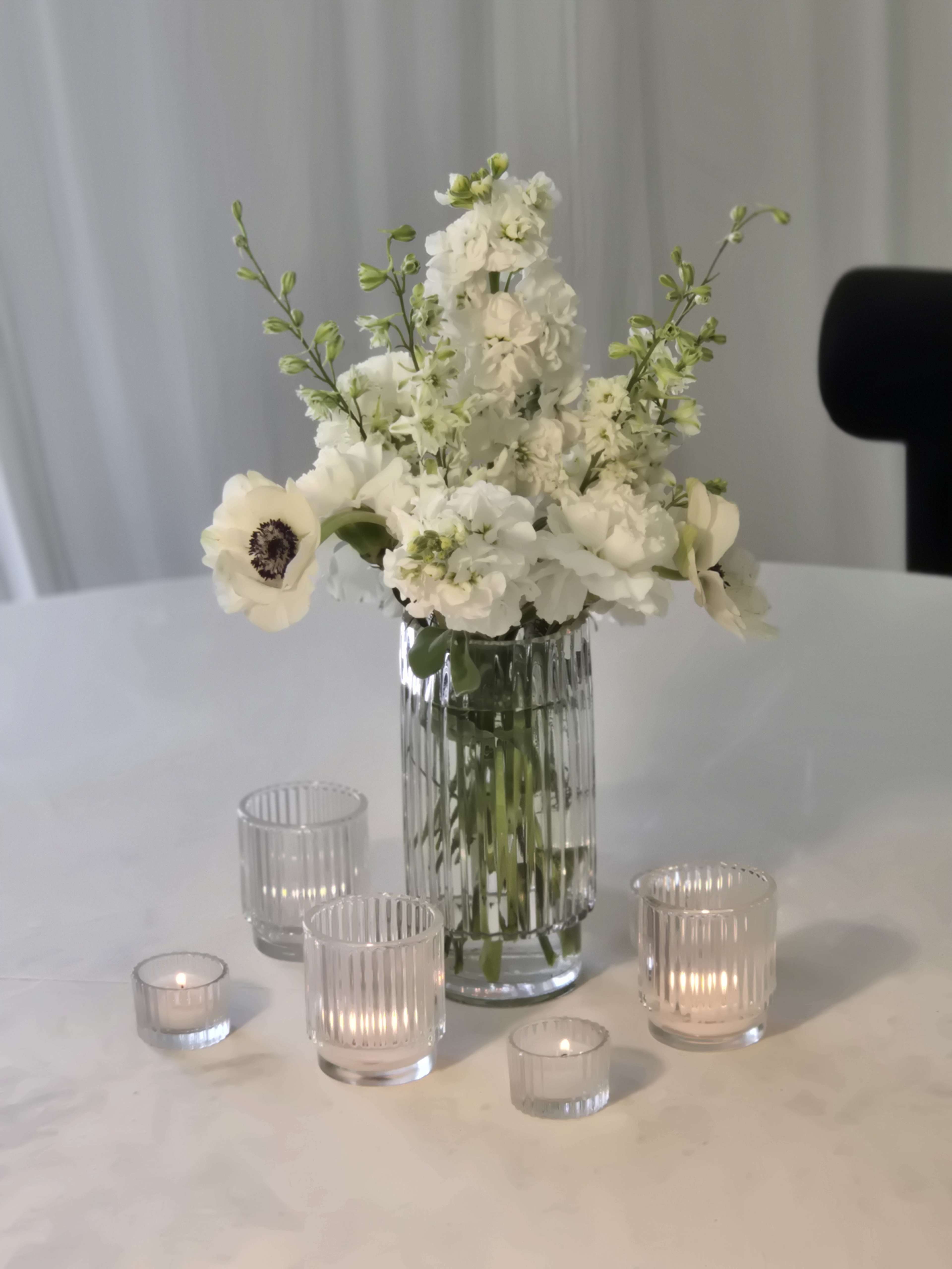 A clear glass vase filled with white flowers is placed on a white table, surrounded by several small glass candle holders.
