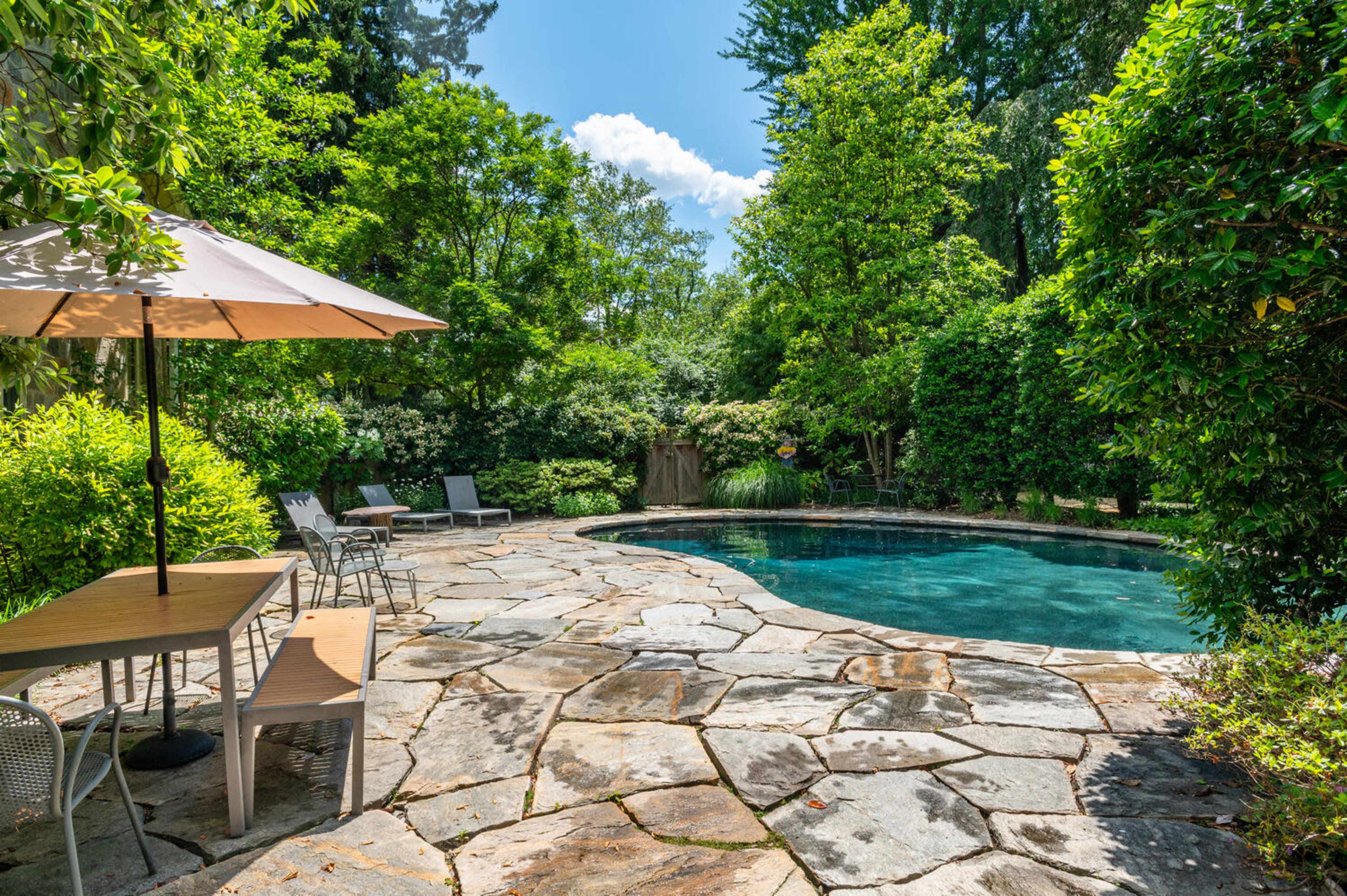 A stone patio surrounds a swimming pool, with lounge chairs and an umbrella nearby, set within a lush garden.