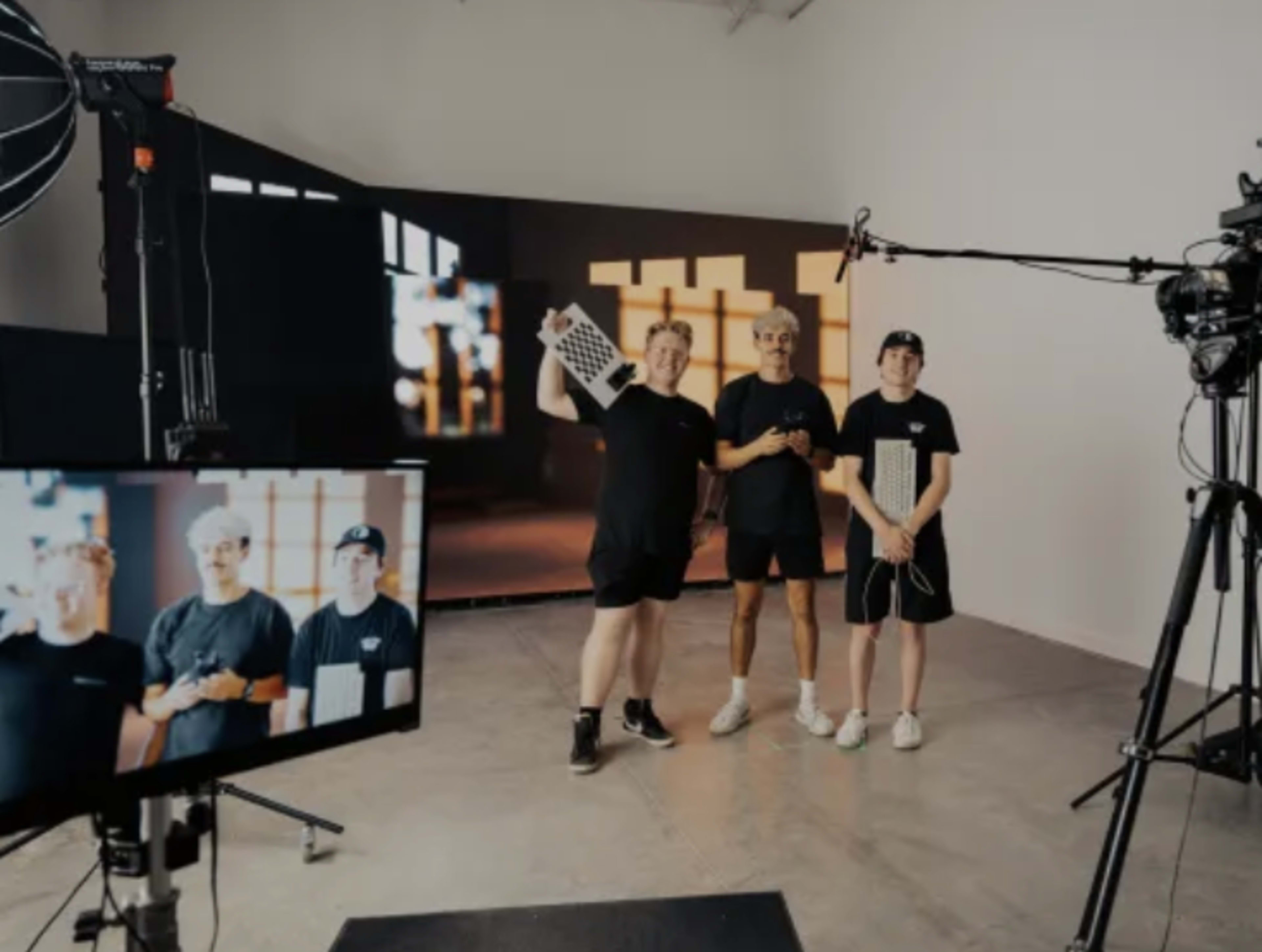 Three individuals stand in a studio set, holding props and surrounded by equipment, with a large screen displaying an abstract design in the background.