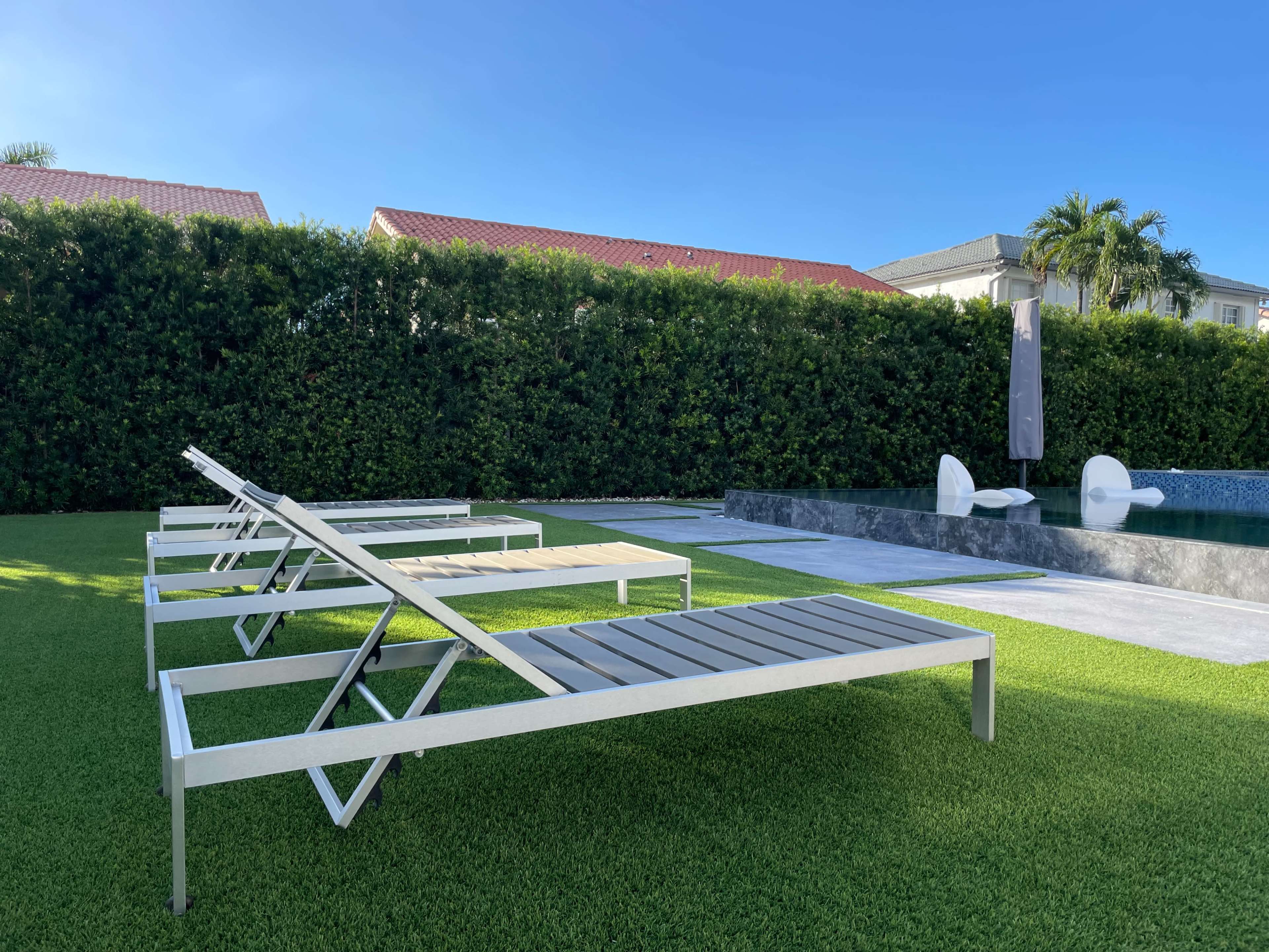 The image shows several lounge chairs arranged beside a swimming pool, set against a backdrop of neatly trimmed hedges and a clear blue sky.
