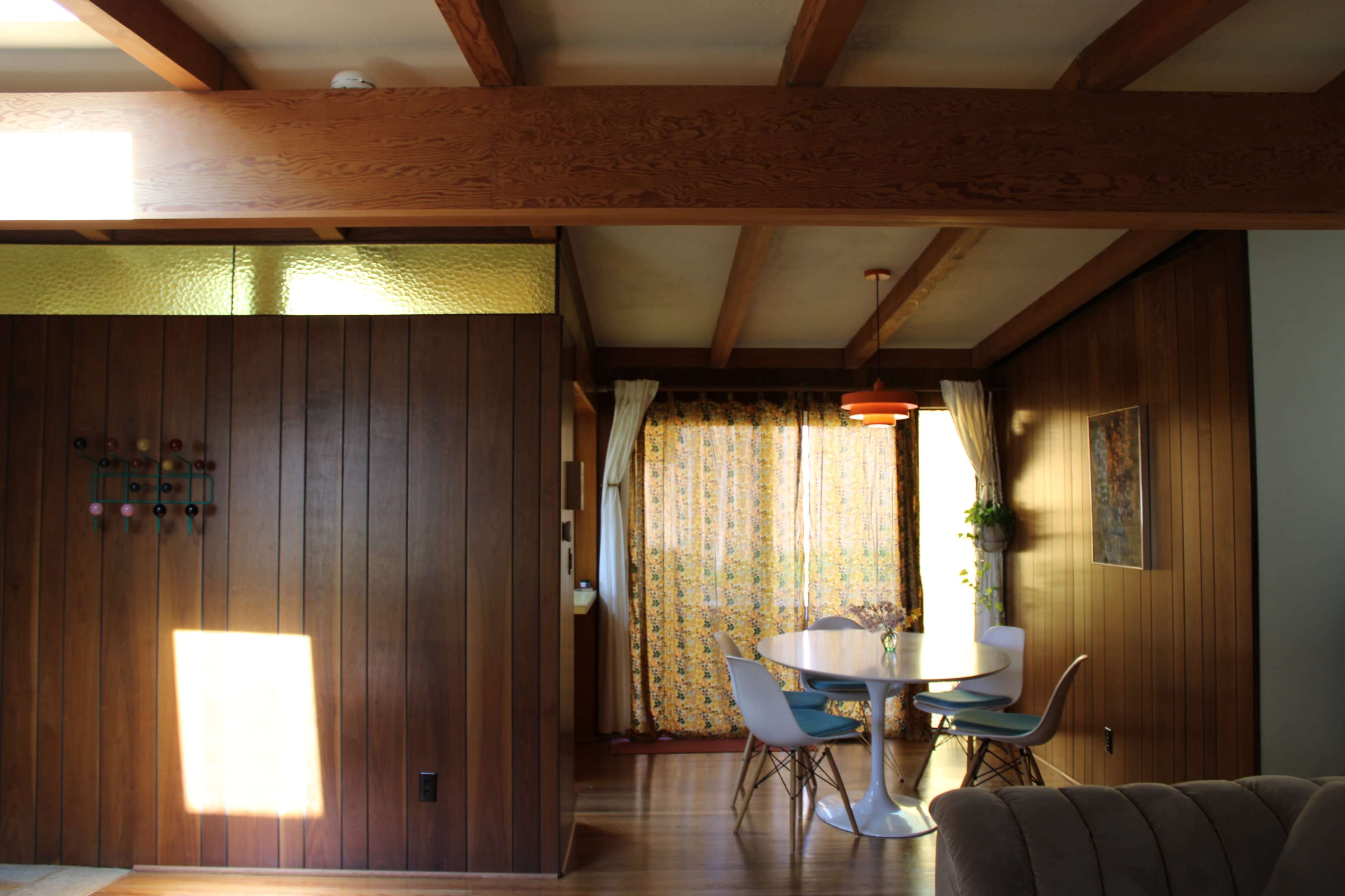 The image shows a mid-century modern interior featuring wood paneling, a round dining table with light blue chairs, and a brightly patterned curtain in the background.
