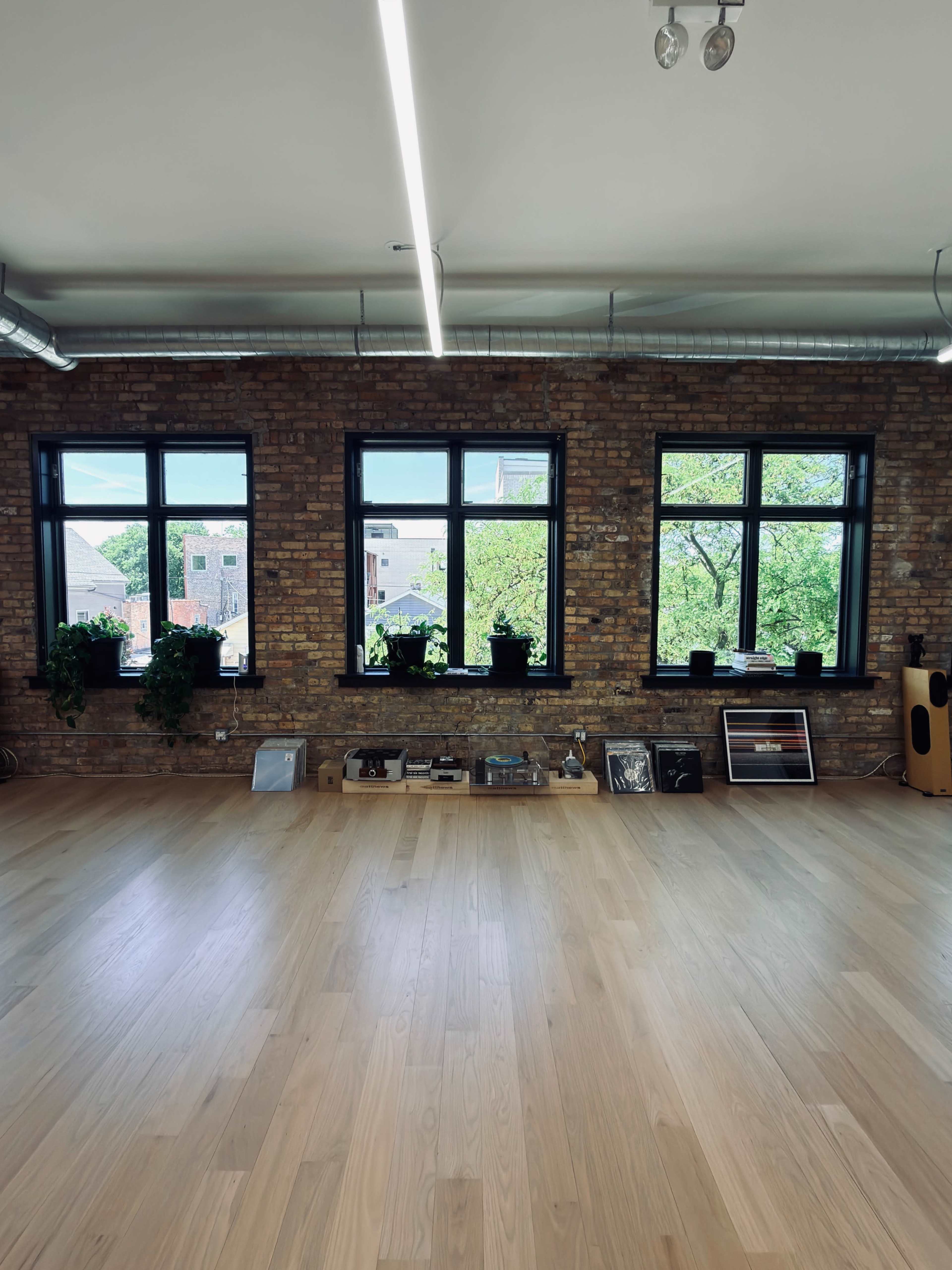The image shows a spacious interior with wooden flooring, exposed brick walls, and large windows adorned with potted plants.