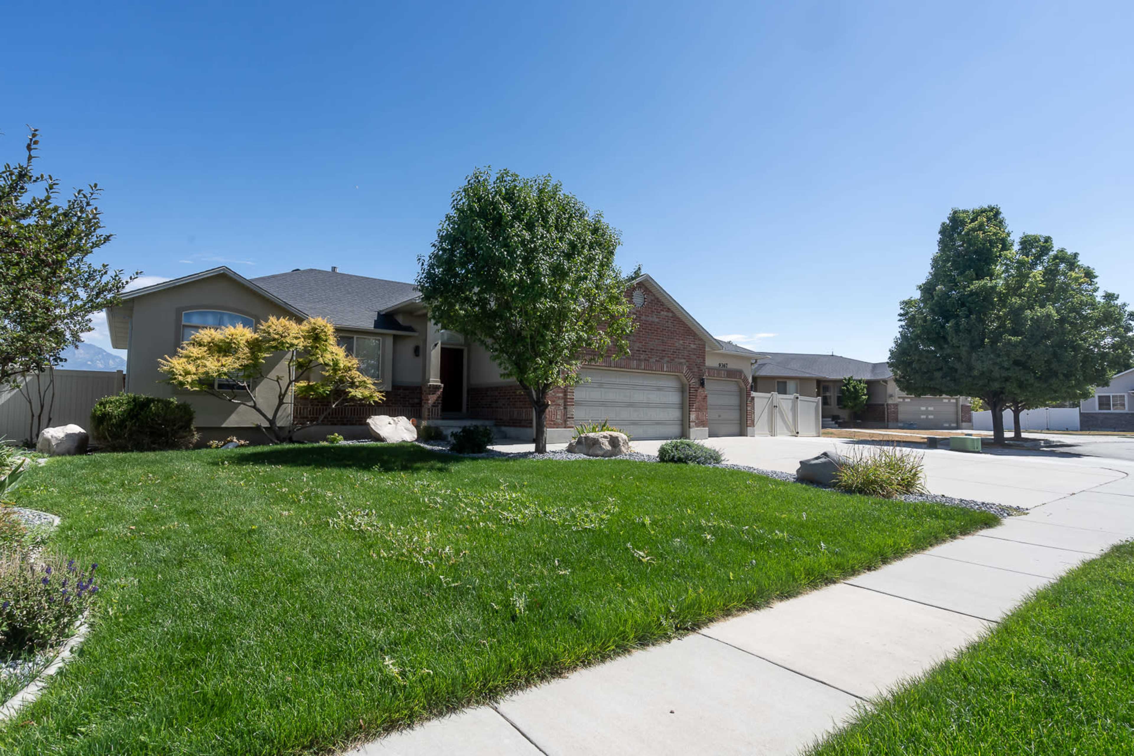 A single-story house with a brick facade and a landscaped yard featuring various trees and shrubs is seen under a clear blue sky.
