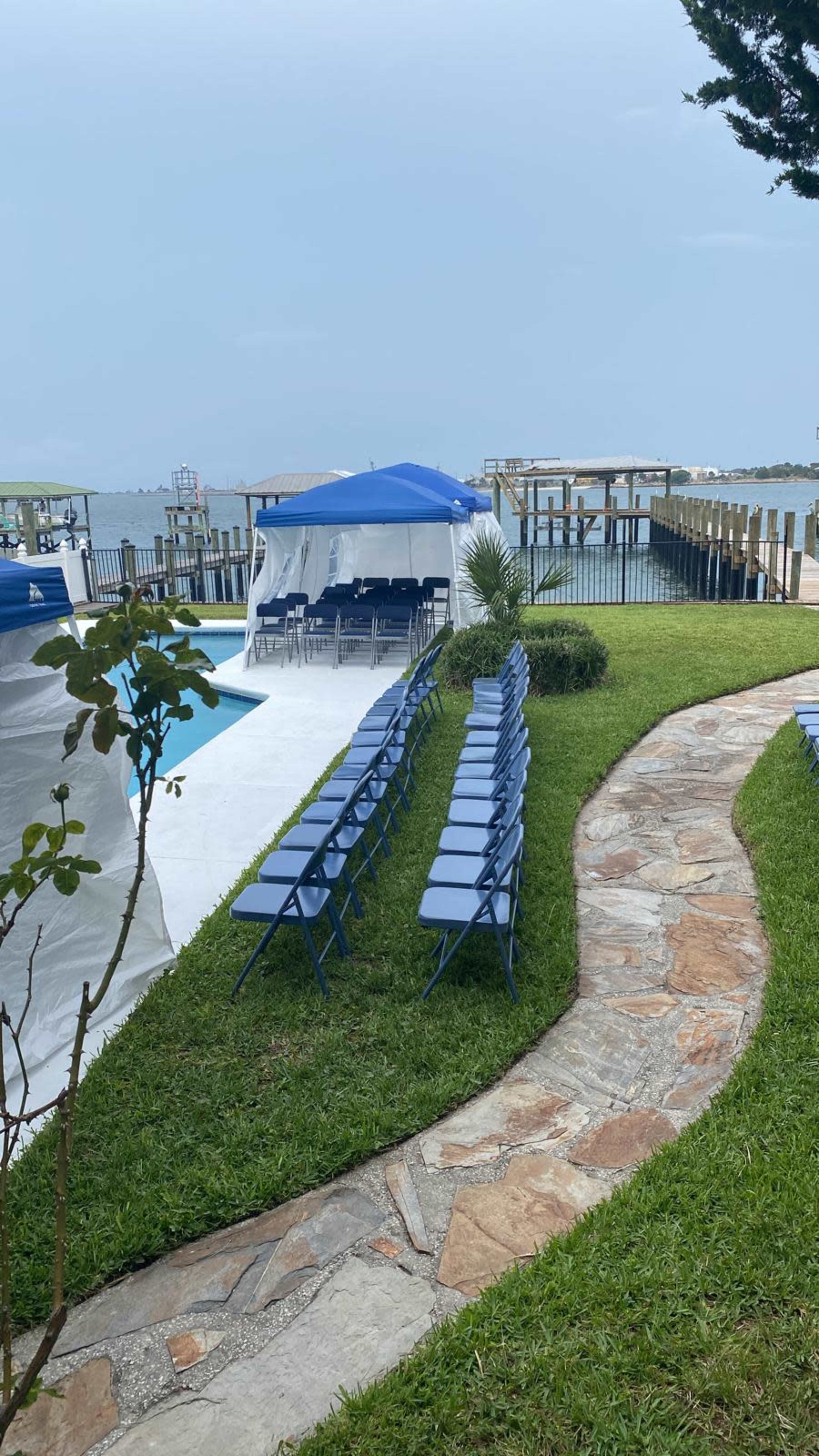The image shows a neat arrangement of blue chairs facing a canopy near a pool, with a pathway leading to a waterfront view.