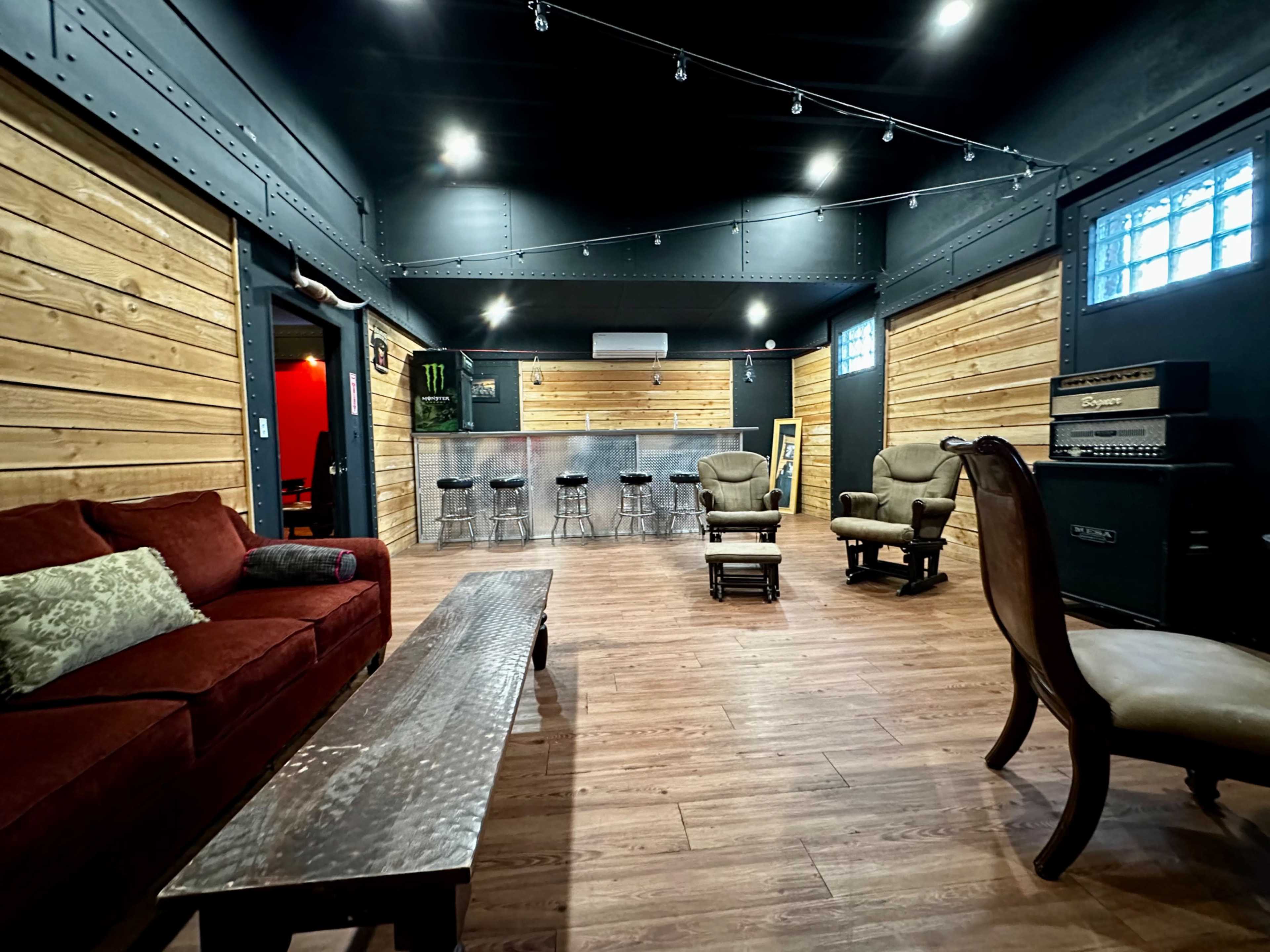 An interior space with wooden walls, a bar area with stools, a red couch, and two armchairs arranged around a coffee table.