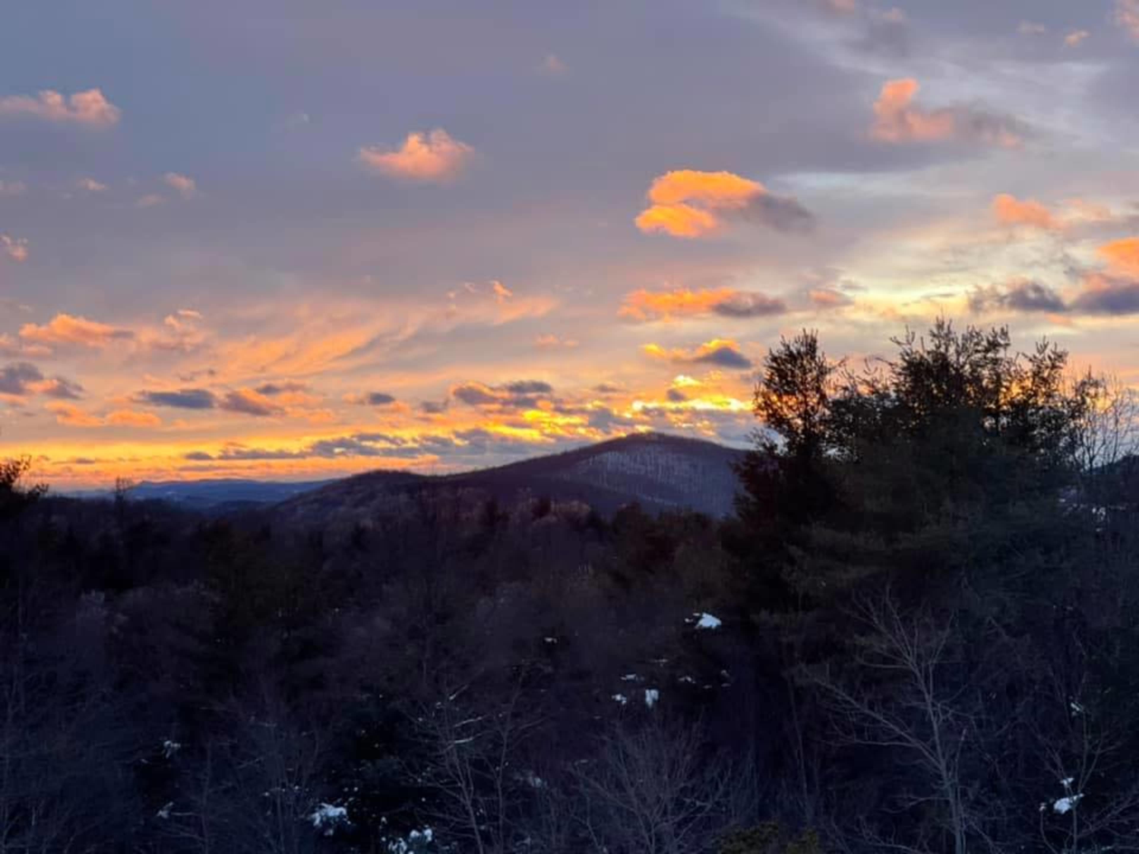 A sunset over a mountain range, with clouds illuminated in shades of orange and pink against a backdrop of trees.