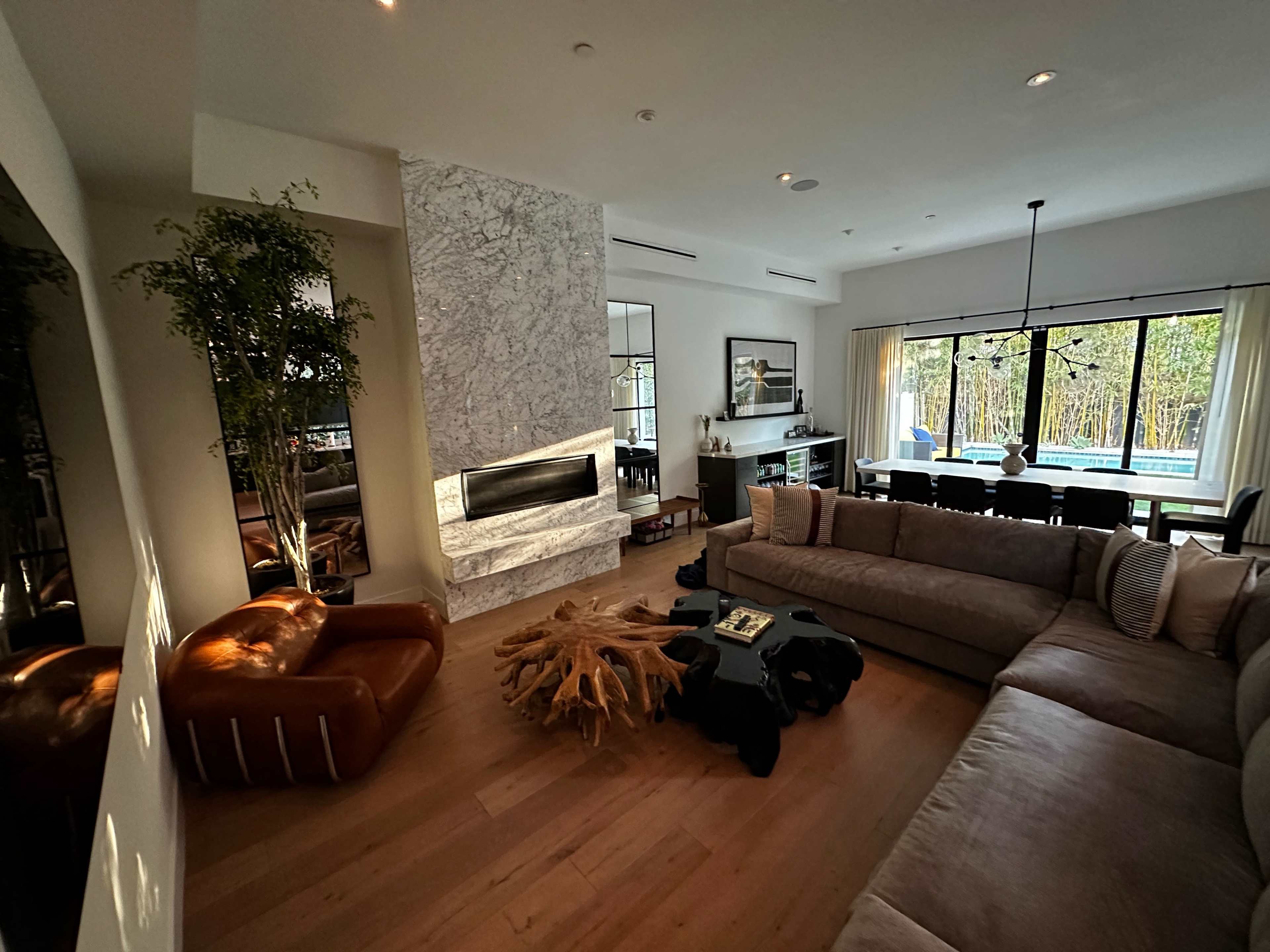 The living room features a large sectional sofa, a coffee table made from wood, and a fireplace surrounded by marble, with large windows overlooking a pool area.