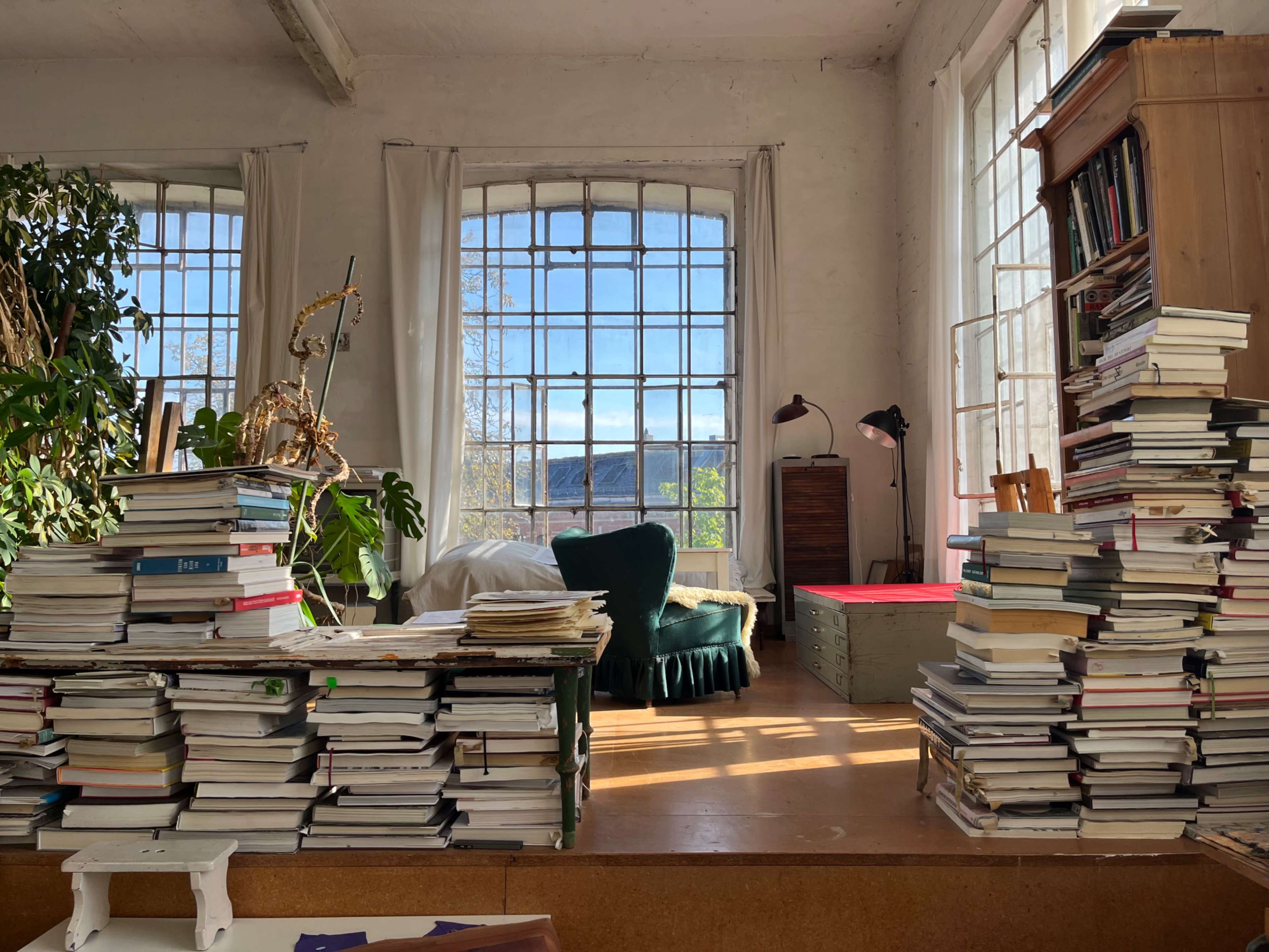 A cluttered room filled with stacked books on both sides, large windows letting in natural light, and a green armchair in the corner.