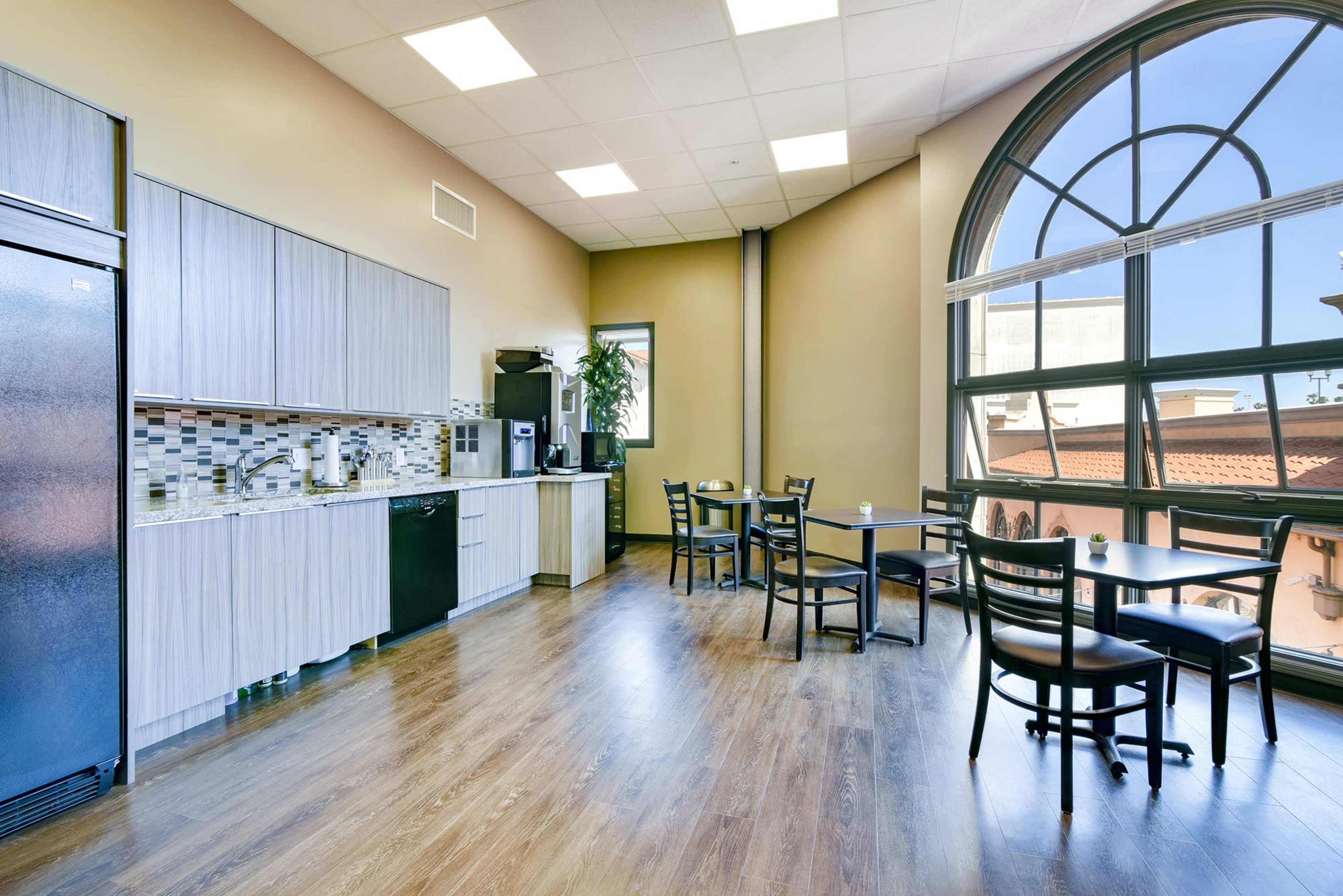 A modern kitchen area features a refrigerator, coffee maker, and seating with large windows that provide natural light.