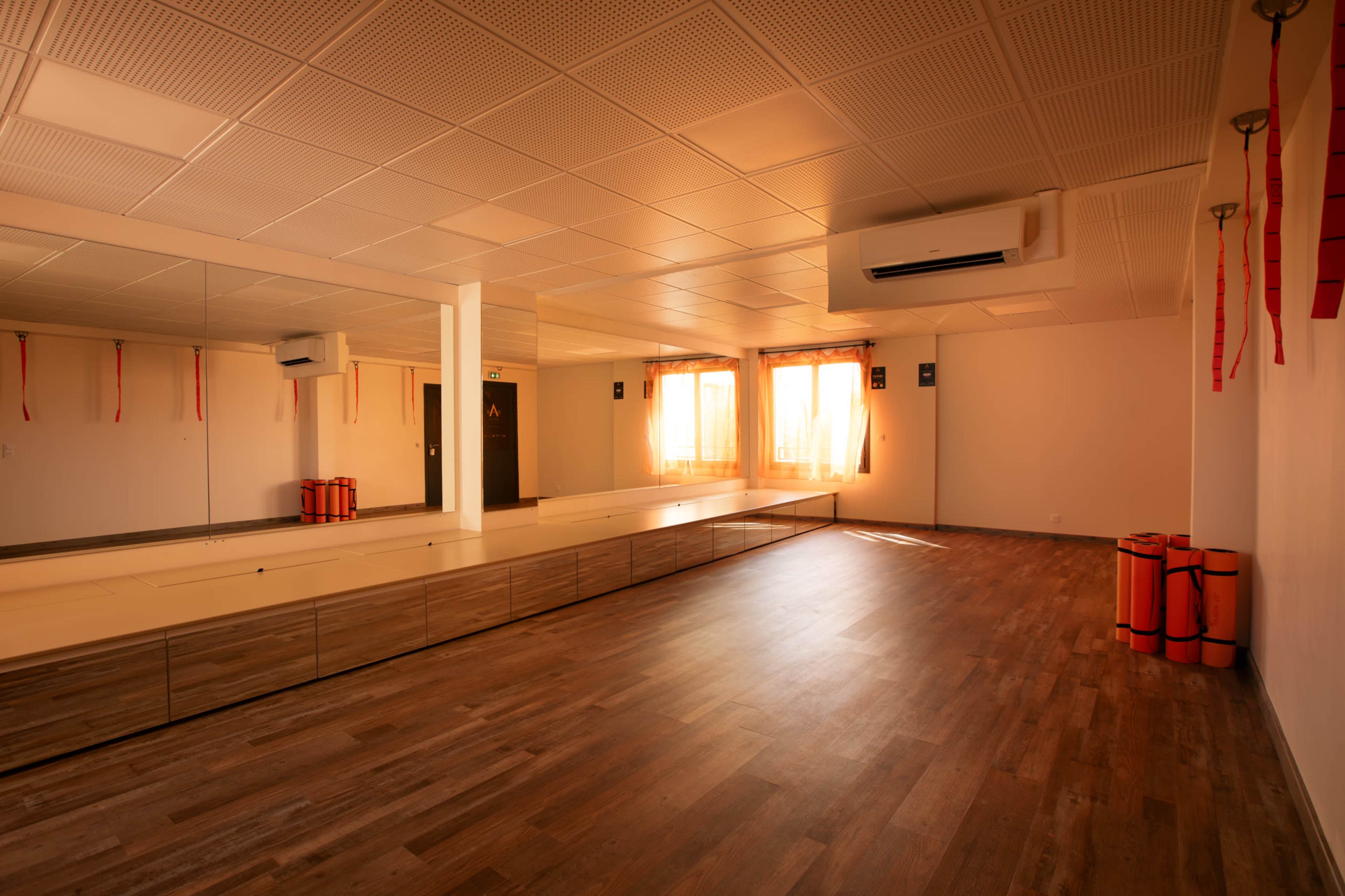 The image shows a well-lit, empty martial arts studio with wooden flooring, mirrors on one wall, and neatly arranged mats.