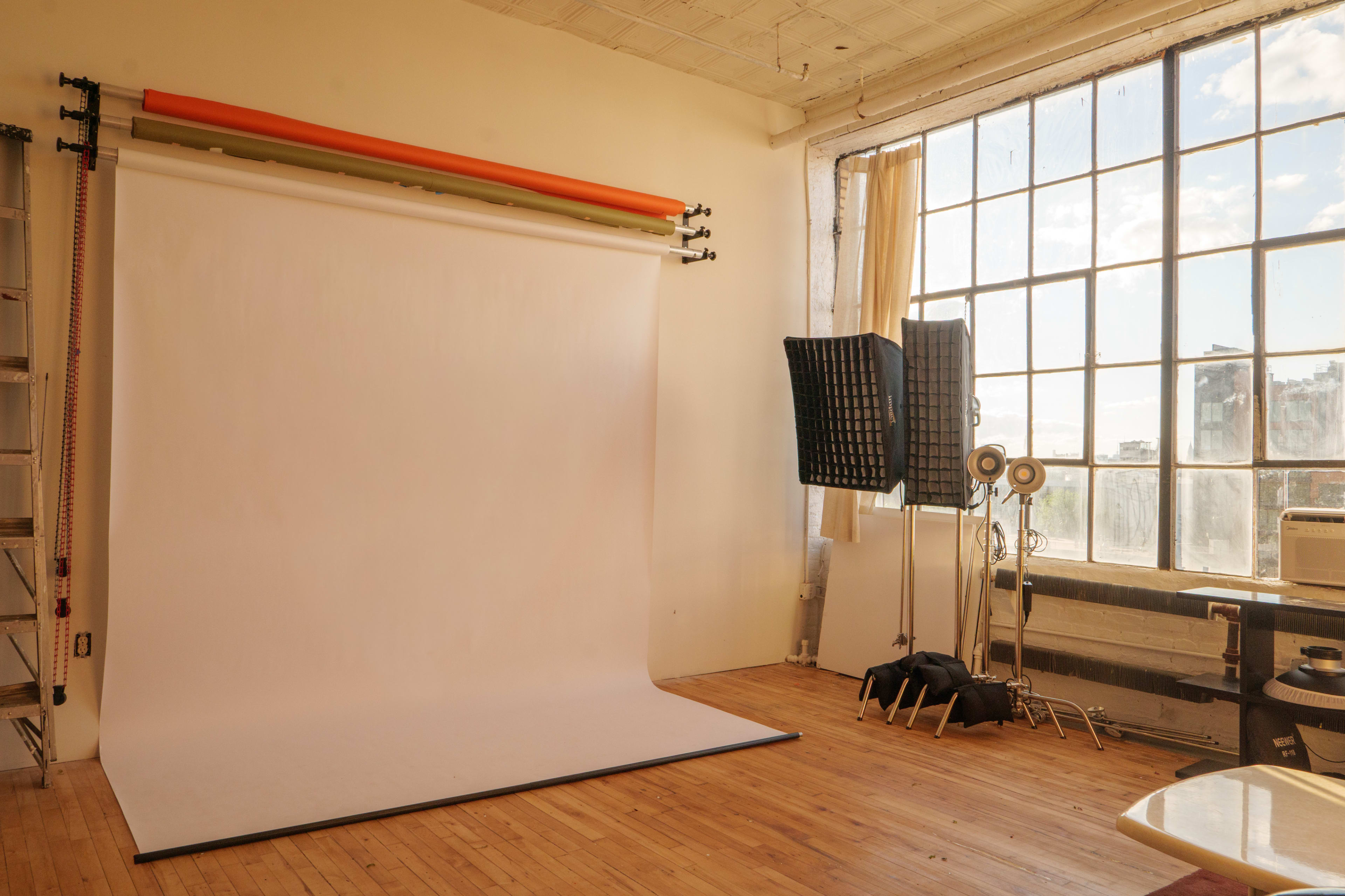 A photography studio features a large white backdrop, natural light streaming through a large window, and various lighting equipment.