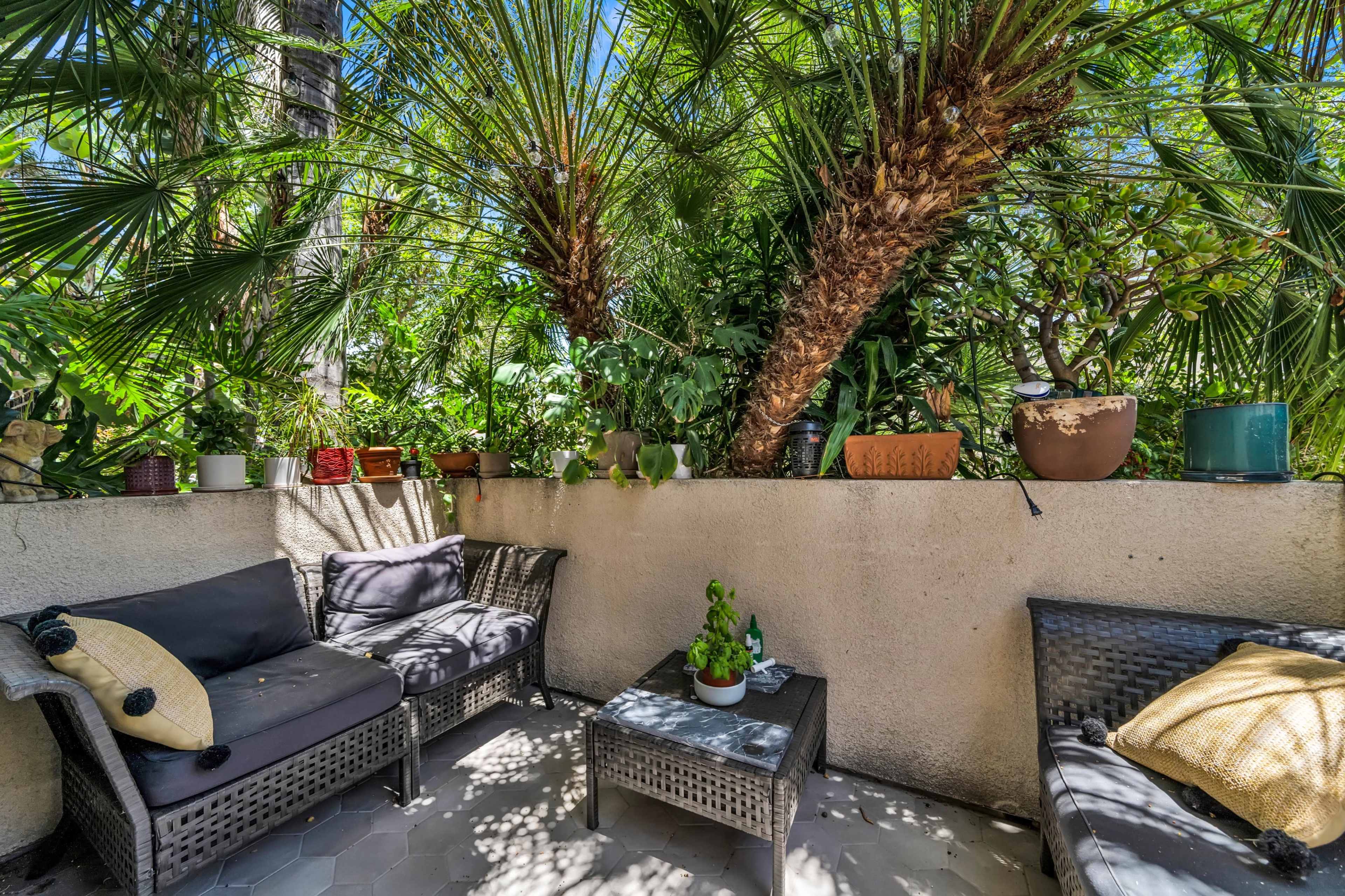 A small outdoor seating area features wicker furniture surrounded by lush greenery and potted plants against a stone wall.