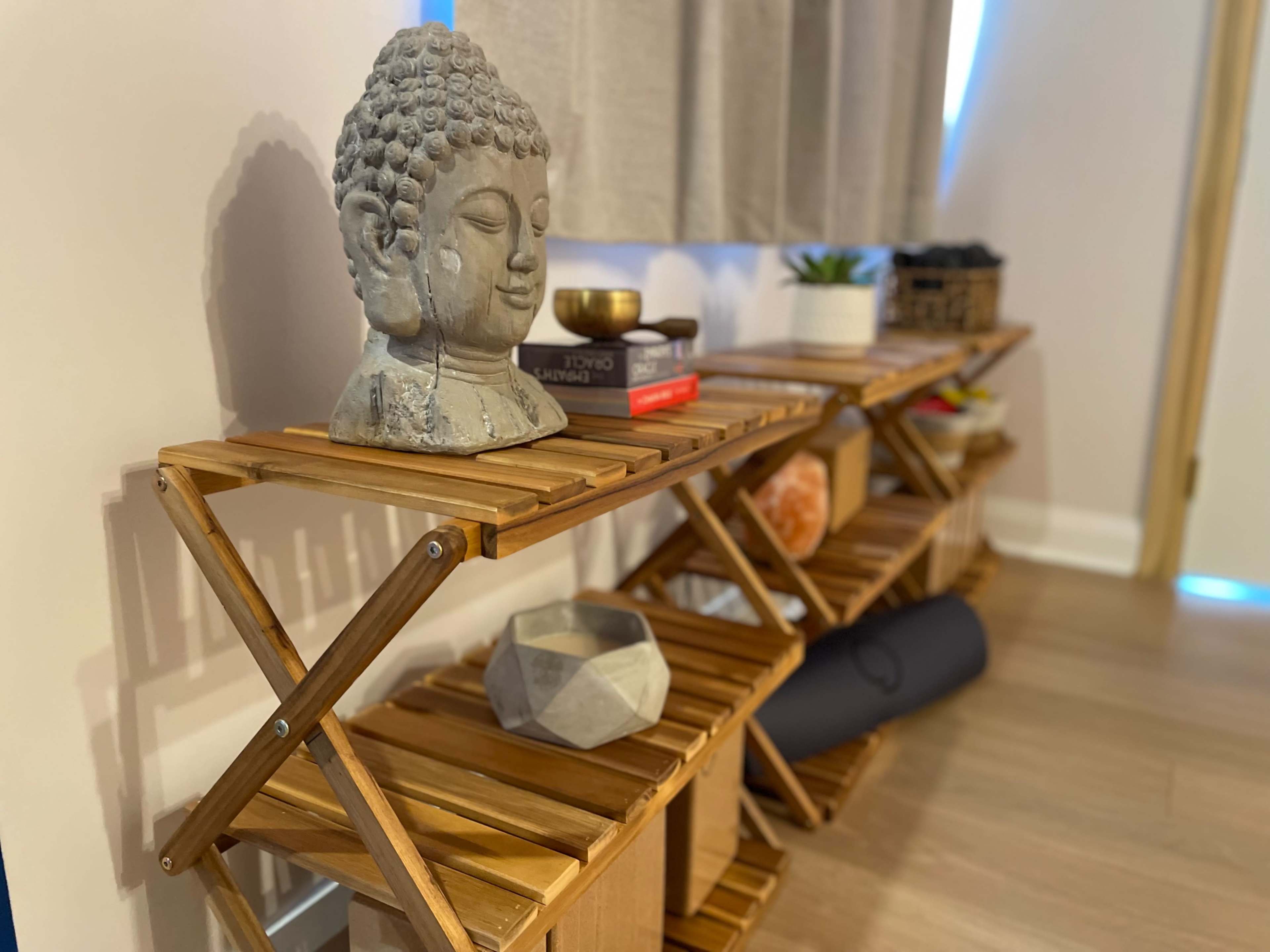 A wooden shelf unit with three tiers, featuring a Buddha statue, a small bowl, books, and a yoga mat.