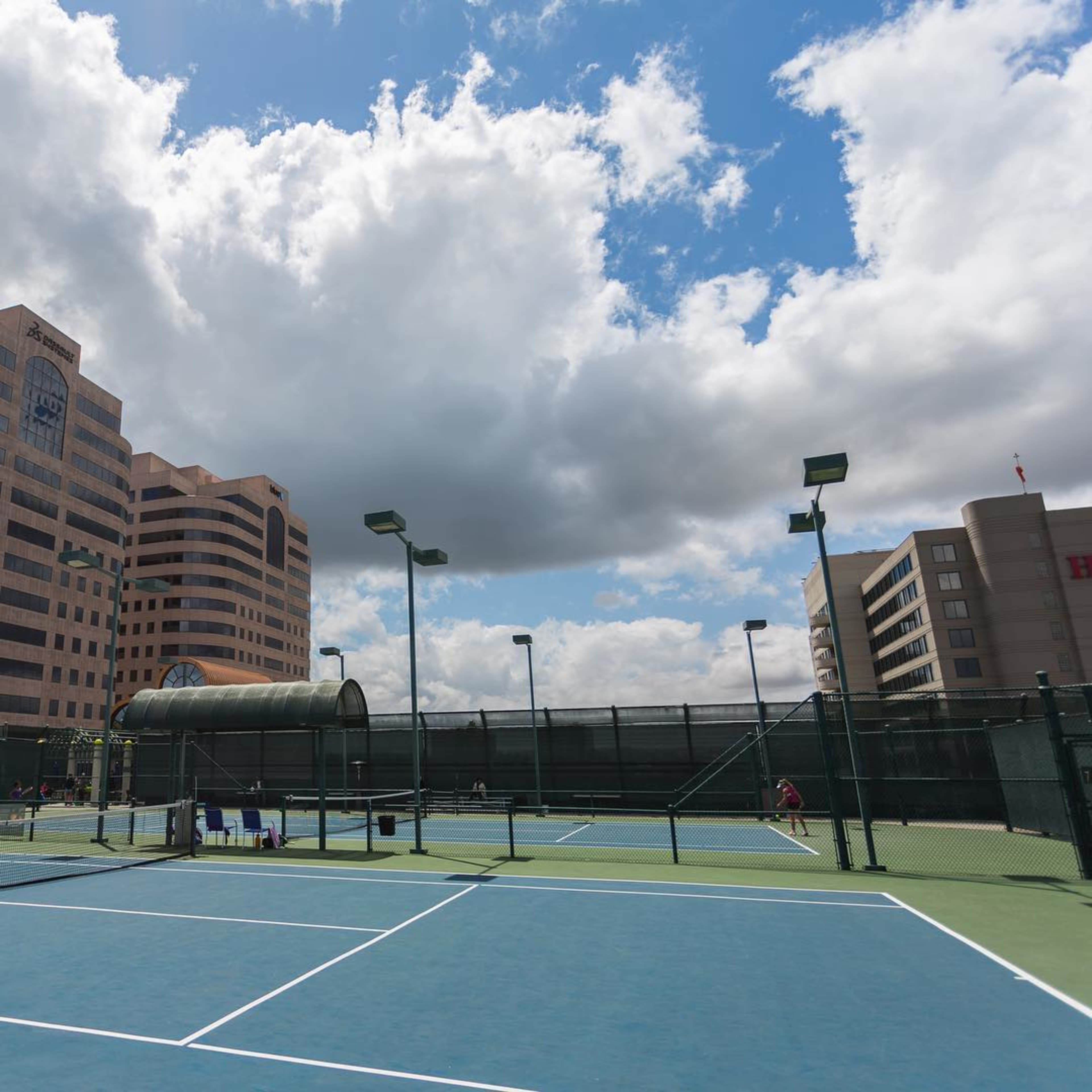 A tennis court surrounded by buildings and partly cloudy skies.